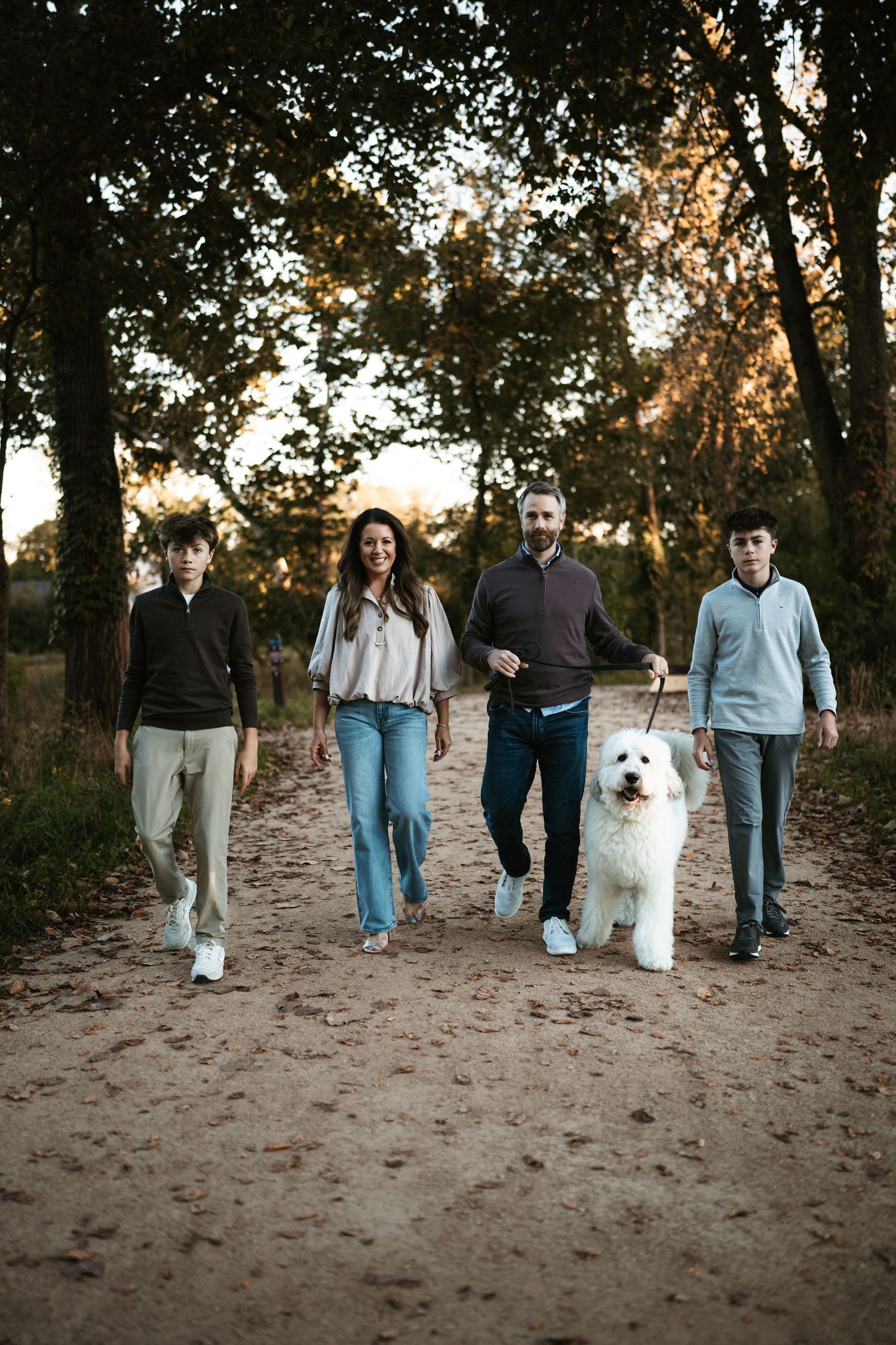Family of four walking a large white fluffy dog on a dirt path through autumn woods at golden hour.