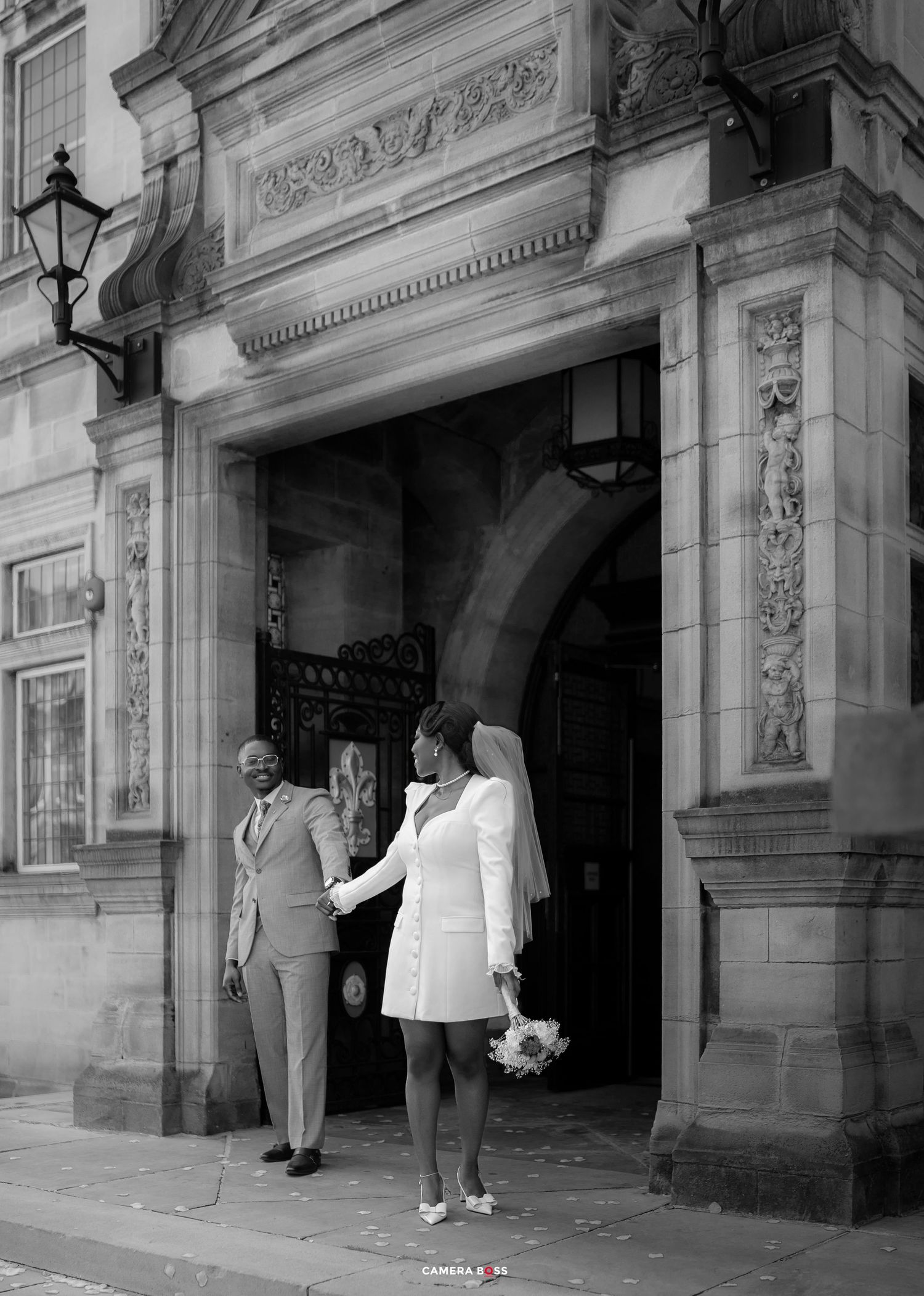 Black and white photo of a couple holding hands at an ornate stone building entrance, bride in short white dress with veil and bouquet.