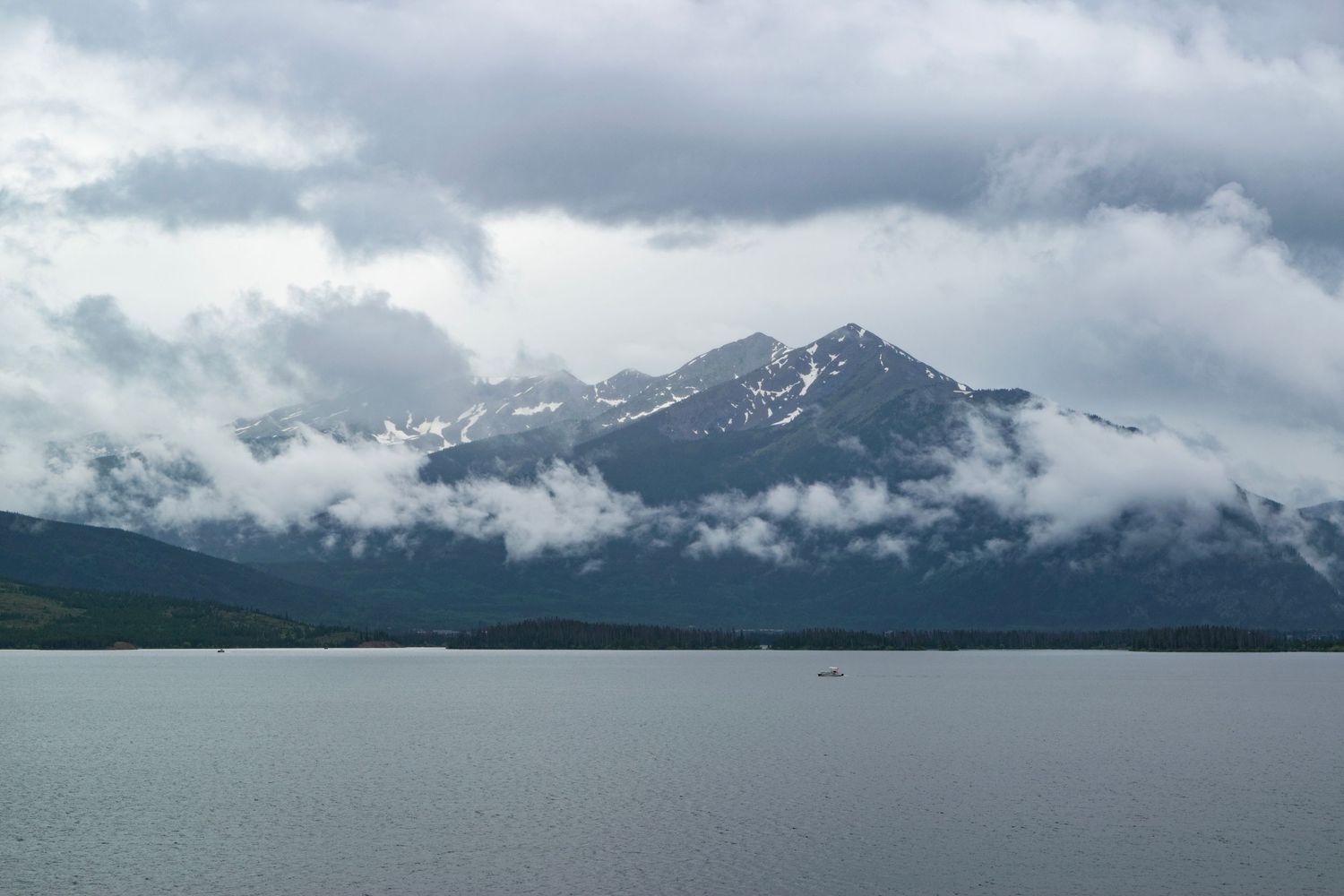 Misty mountain peaks rise above a tranquil lake with low-hanging clouds in dramatic weather.