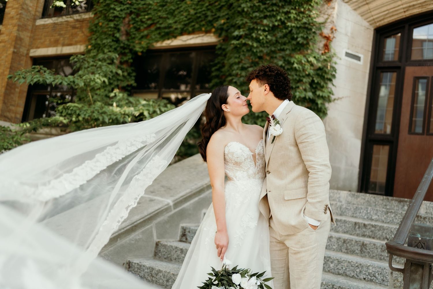 A romantic wedding portrait with a flowing veil against an ivy-covered building.