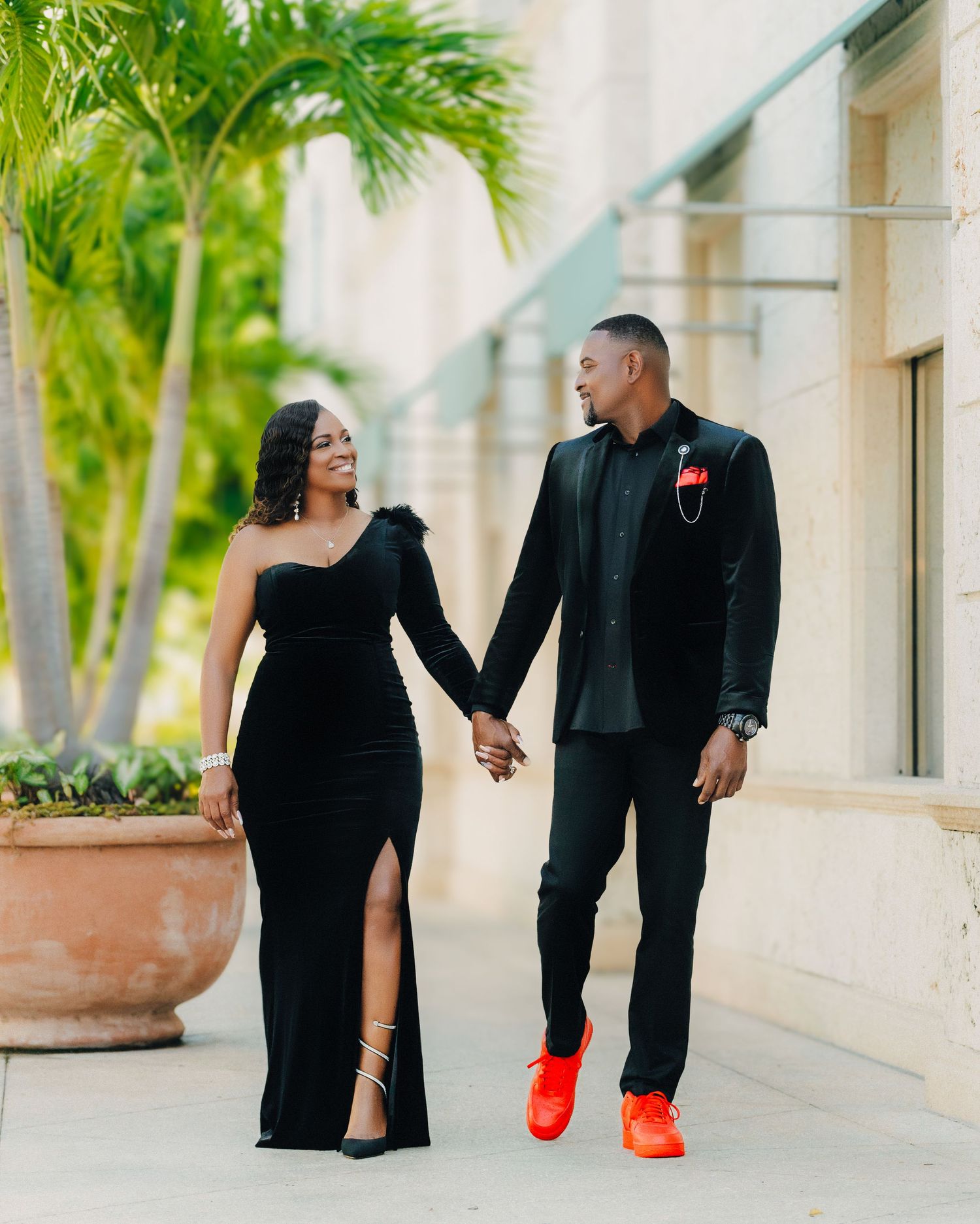 Couple in formal black attire holding hands while walking along palm-lined path.