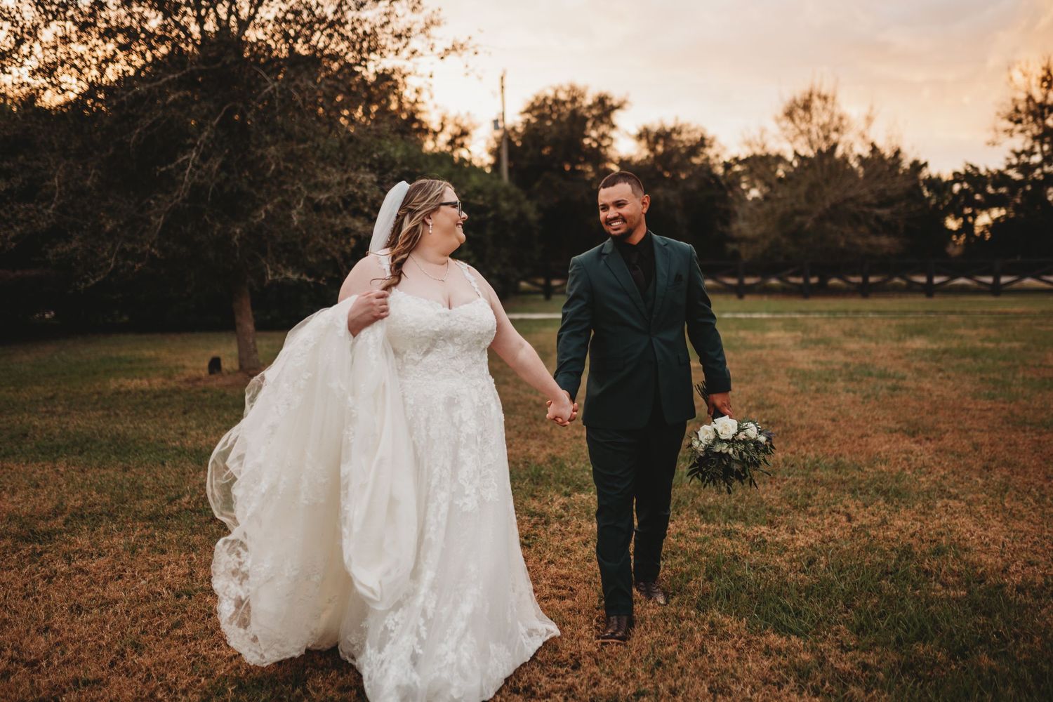 Couple walking hand in hand through a rustic field at sunset in wedding attire.