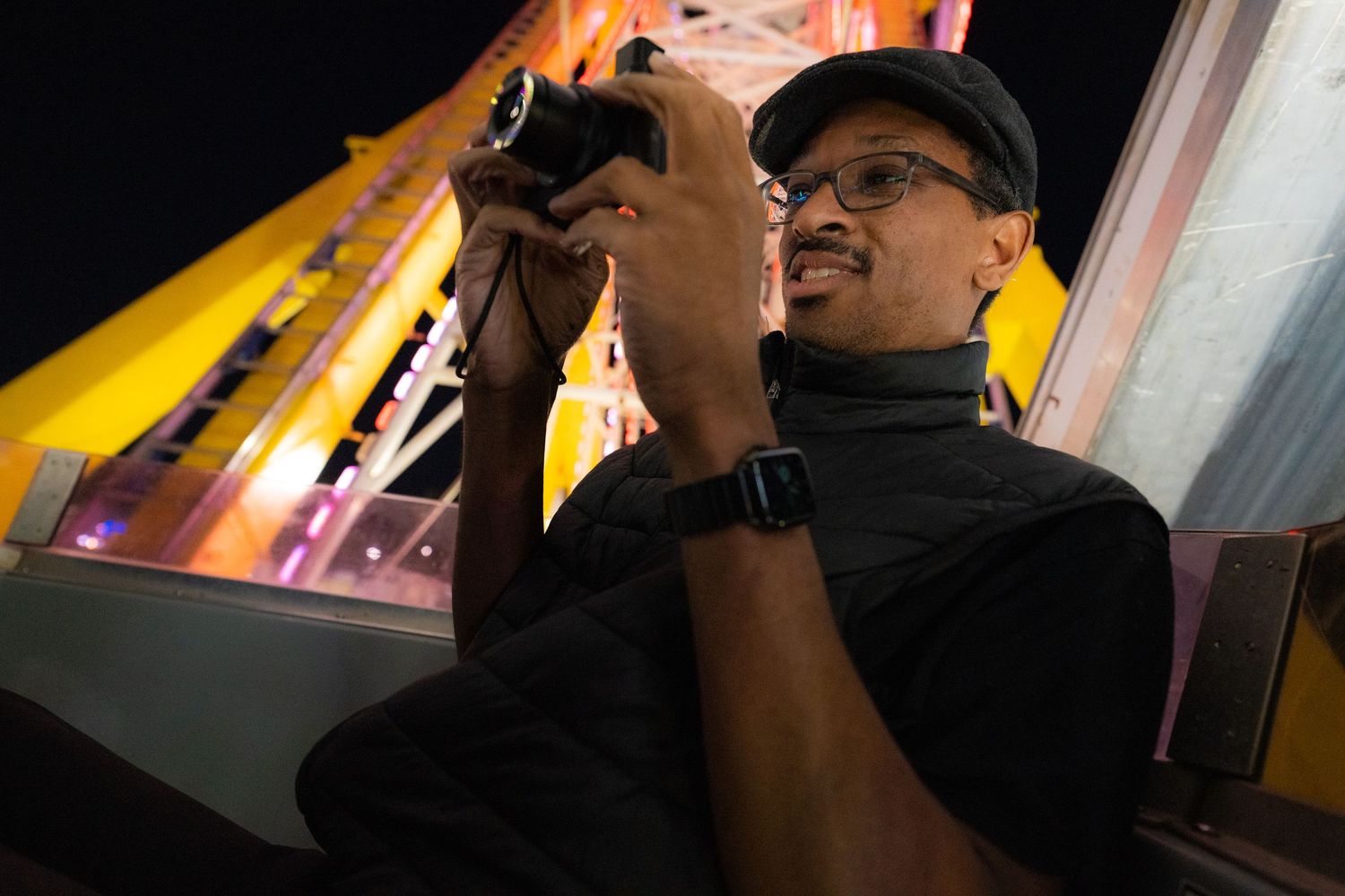 Black man with mustache and glasses smiles while holding his digital camera. He's riding a ferris wheel at night.