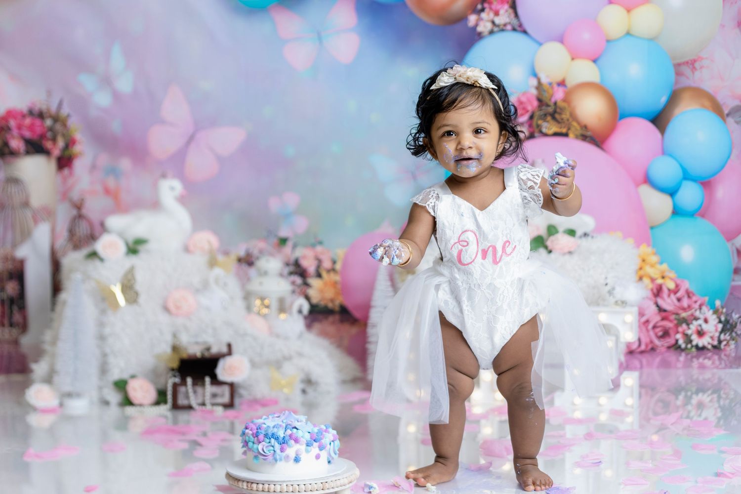 One-year-old girl enjoying a cake smash photoshoot with a pastel balloon and floral backdrop.