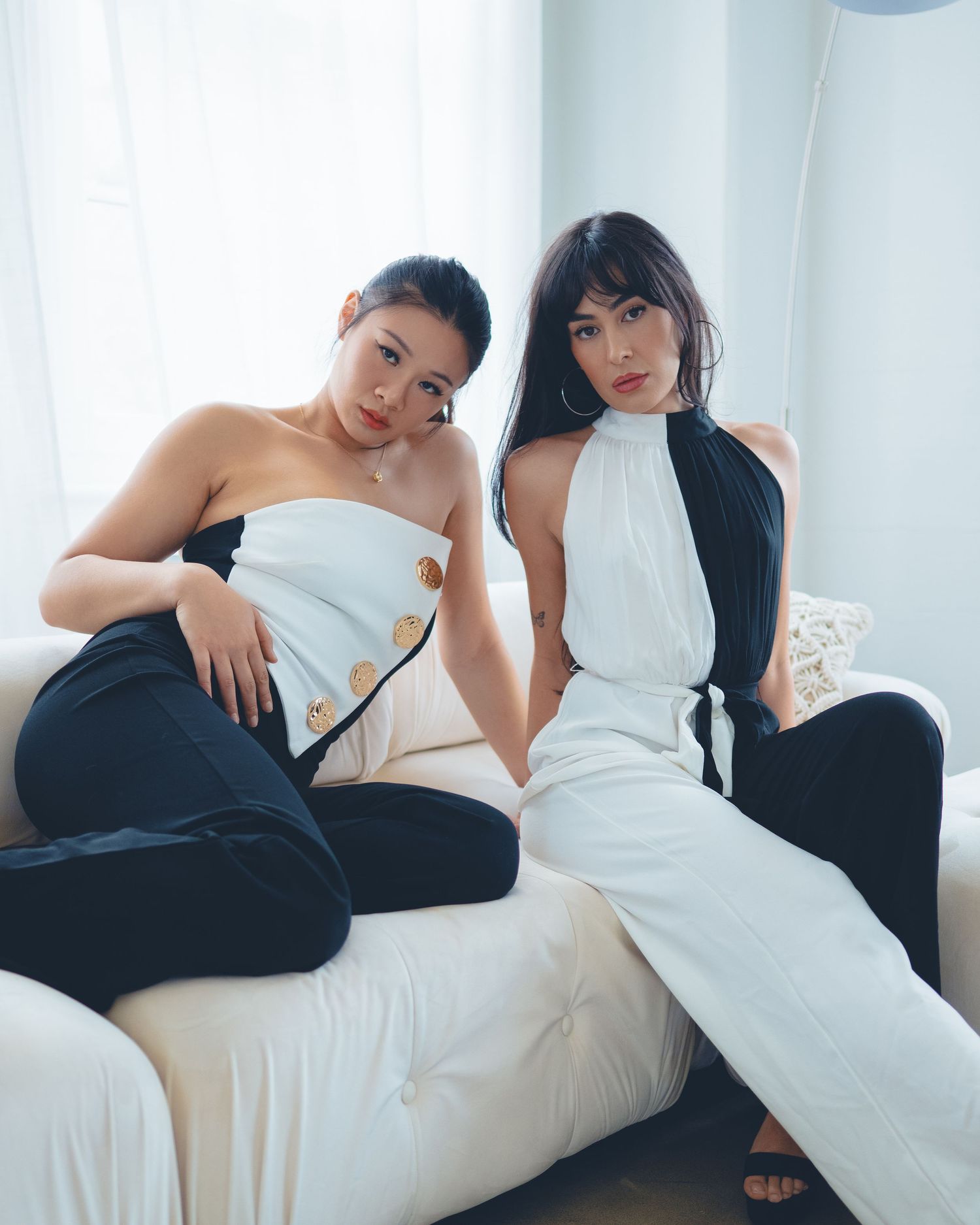 Two fashion models wearing contrasting black and white outfits pose together on a light colored couch in a minimalist setting.