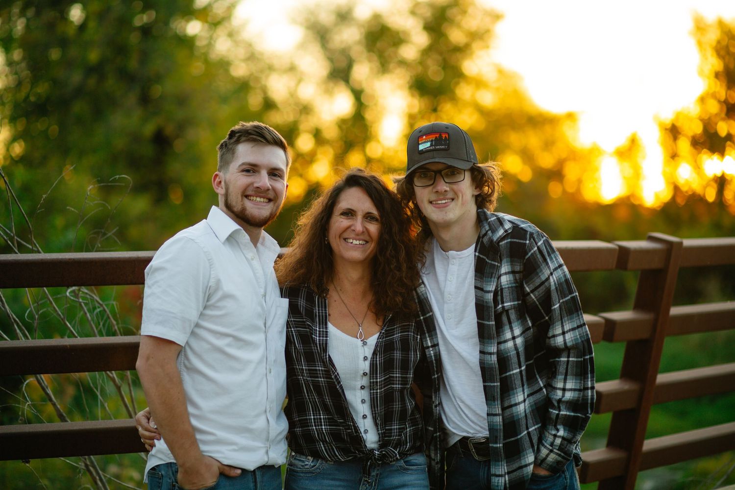 Three people stand smiling together against a wooden fence during golden hour sunset outdoors.