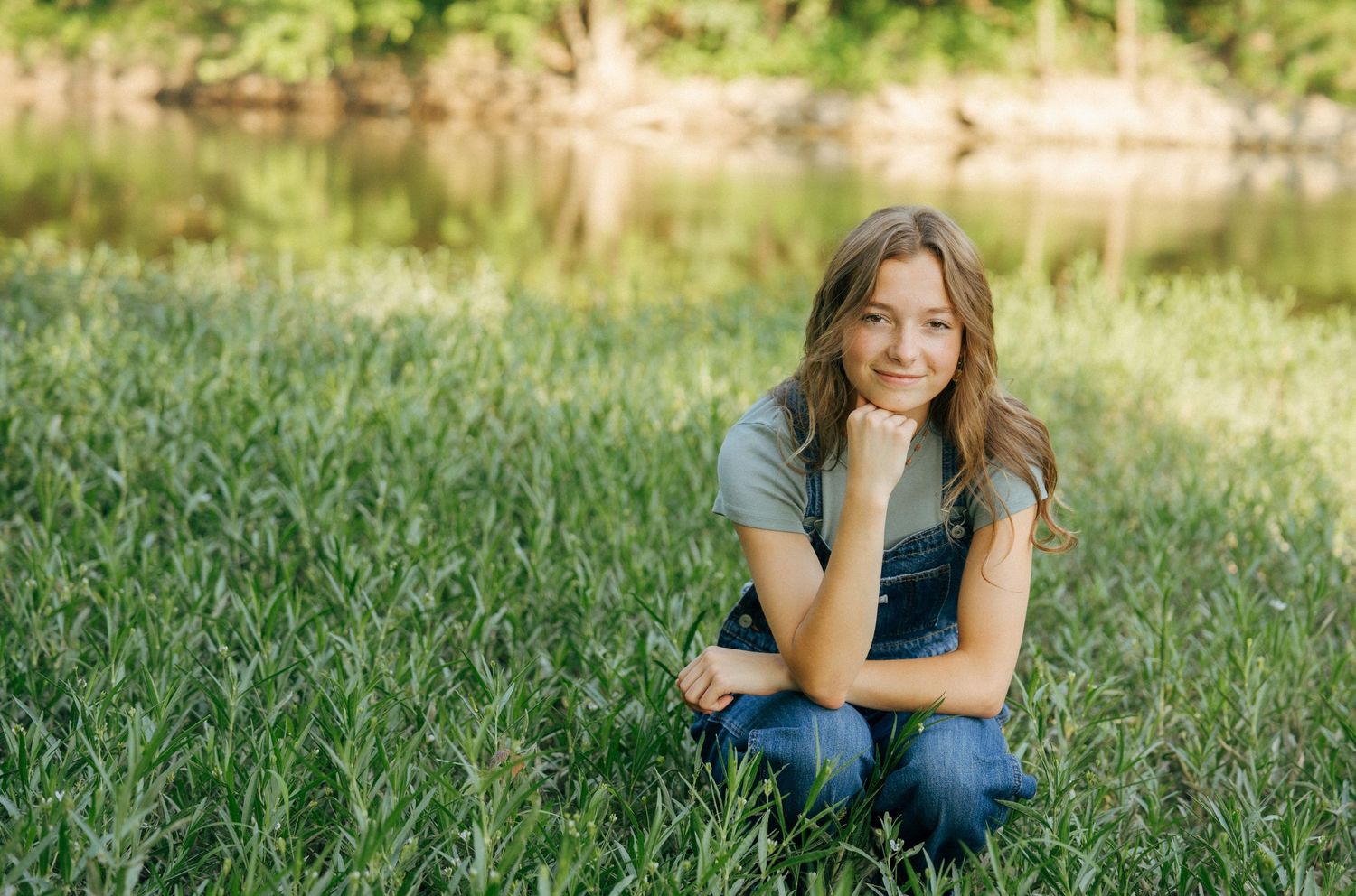 Person in casual attire sitting on grass near a pond on a sunny afternoon.