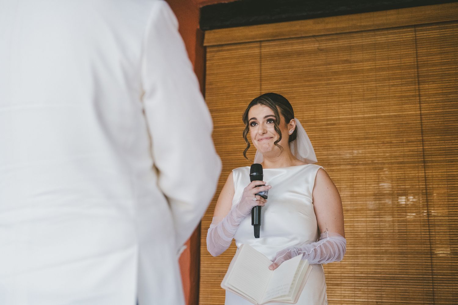 A bride in a white dress and long gloves holds a microphone while speaking during a wedding ceremony.