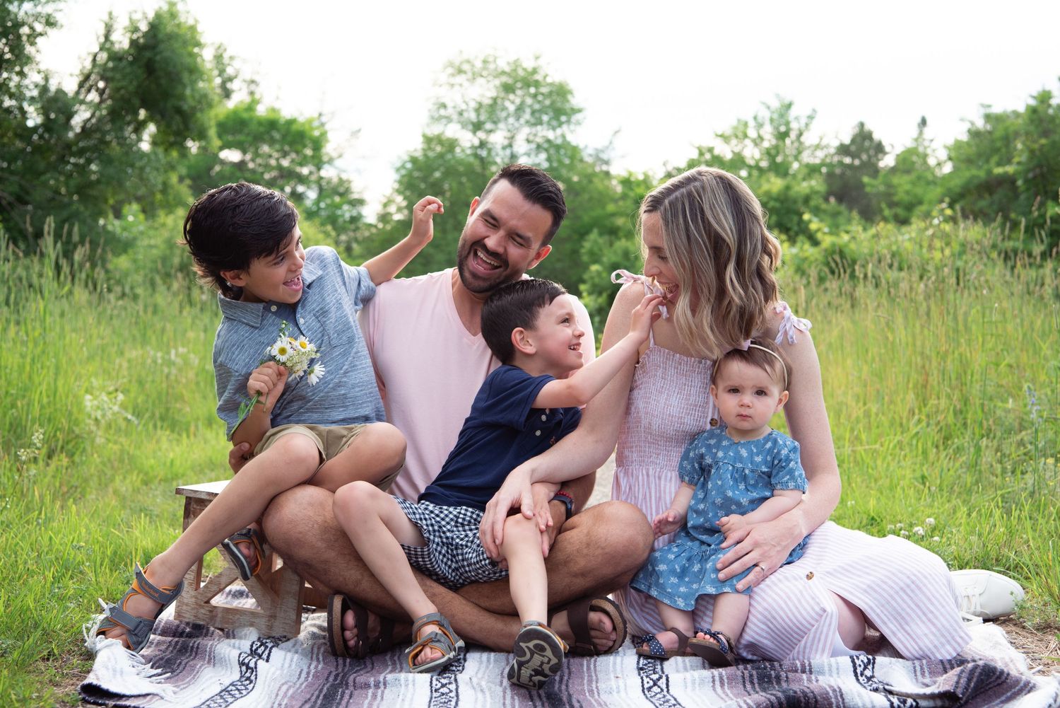 A family enjoying quality time together while sitting on a blanket in a sunny meadow with tall grass and greenery.