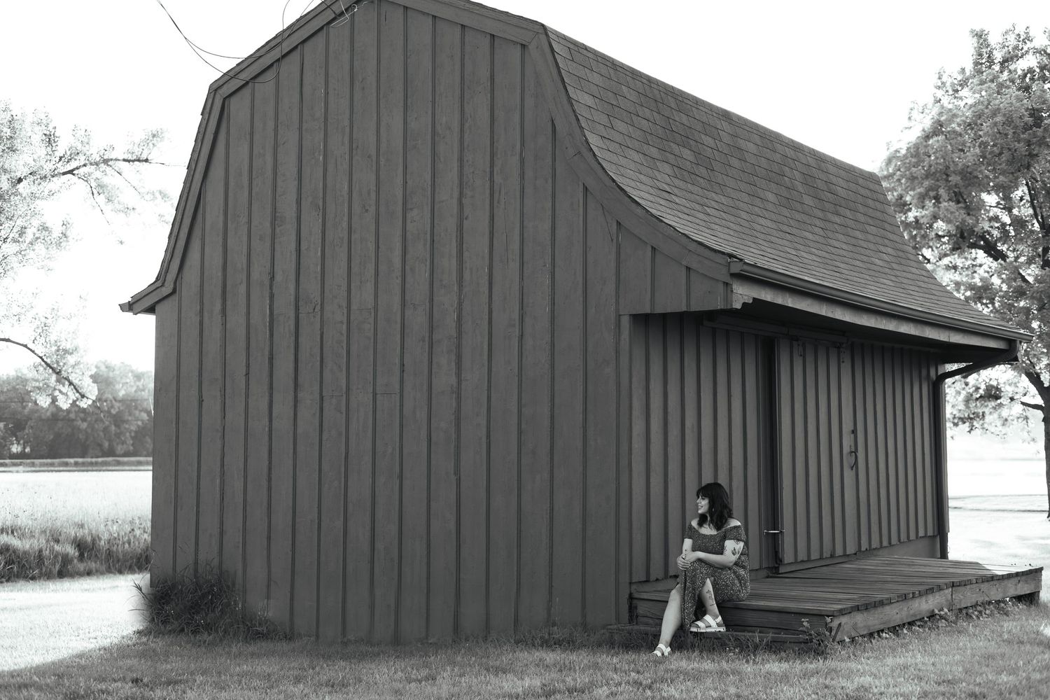 Black and white photograph of a simple wooden barn structure with someone sitting on the porch.