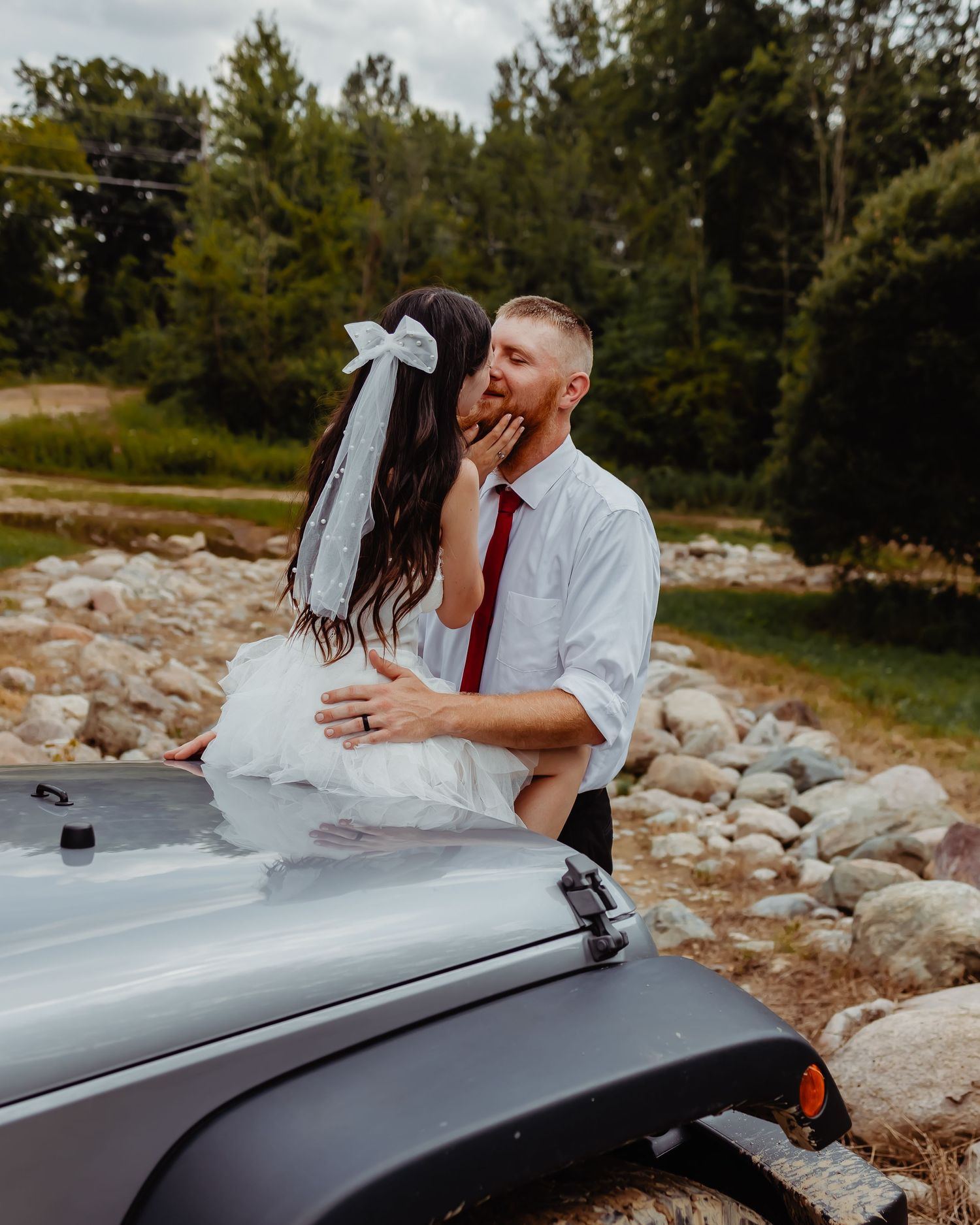 Couple in wedding attire share romantic moment on hood of Jeep with rustic backdrop.