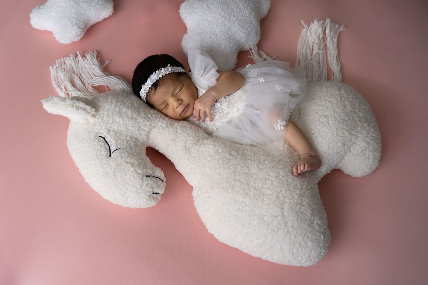 Newborn baby rests peacefully on soft white plush bunny toys against pink background during photo session.