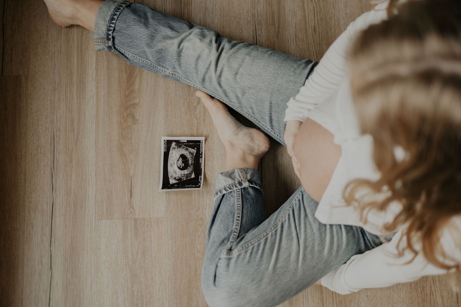 A person lies on a wooden floor looking at an ultrasound sonogram photo placed on the ground.