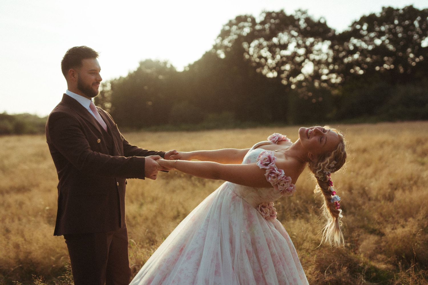 A couple in wedding attire dance together in a golden field at sunset, with the bride dipping backwards gracefully.