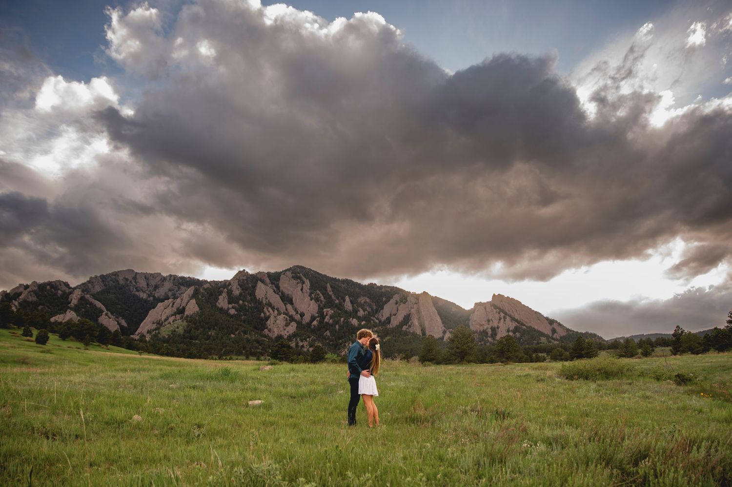 A couple stands together in a meadow beneath dramatic storm clouds and the majestic Flatiron Mountains in Boulder, Colorado.