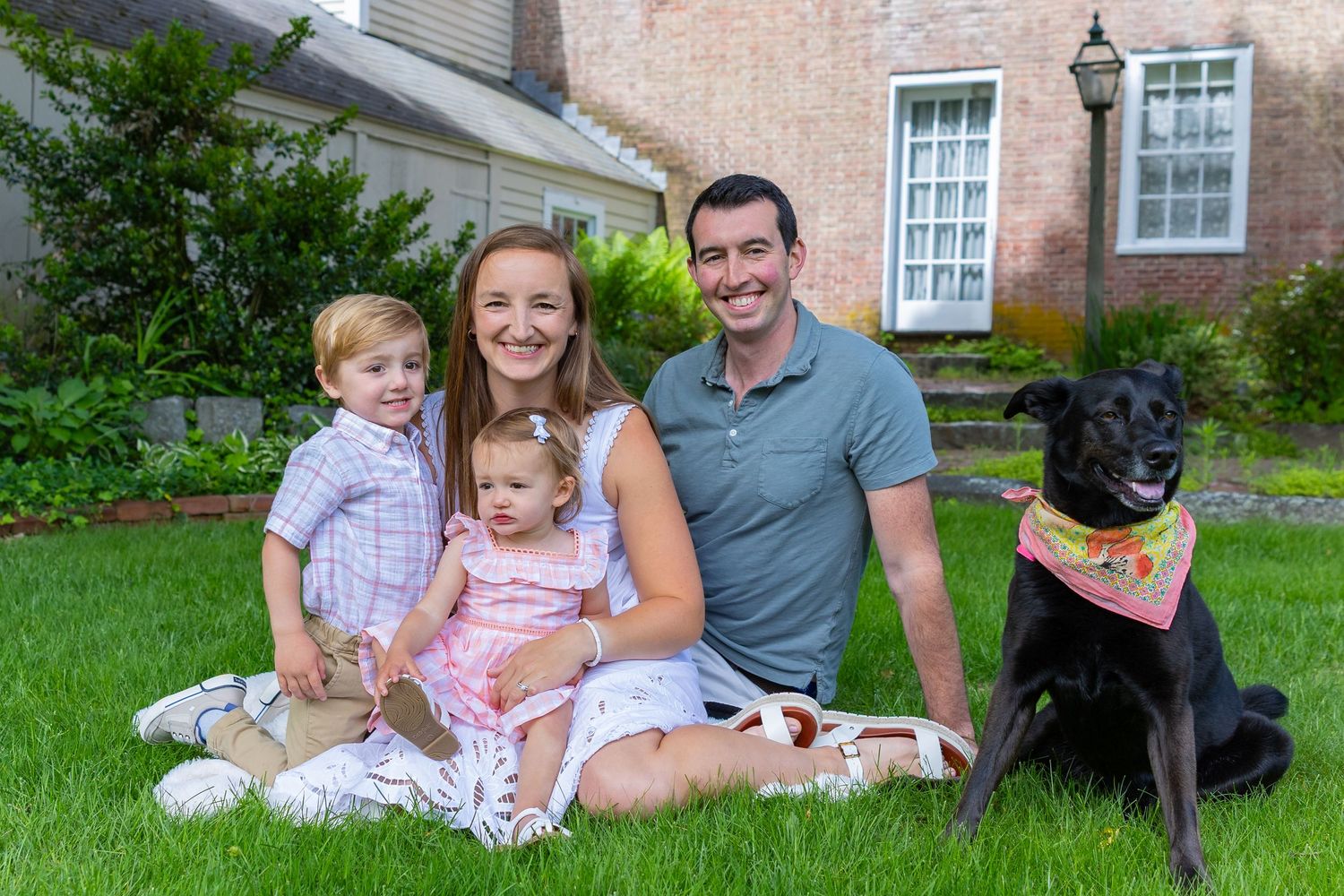 Family with two young children and black dog sitting together on blanket in front of brick house.