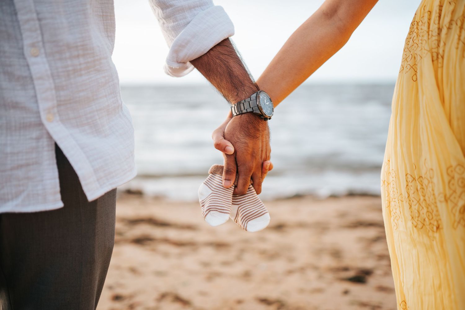 A couple holds hands with newborn socks on a sandy beach with ocean waves in the background taken by Melbourne newborn photographer.