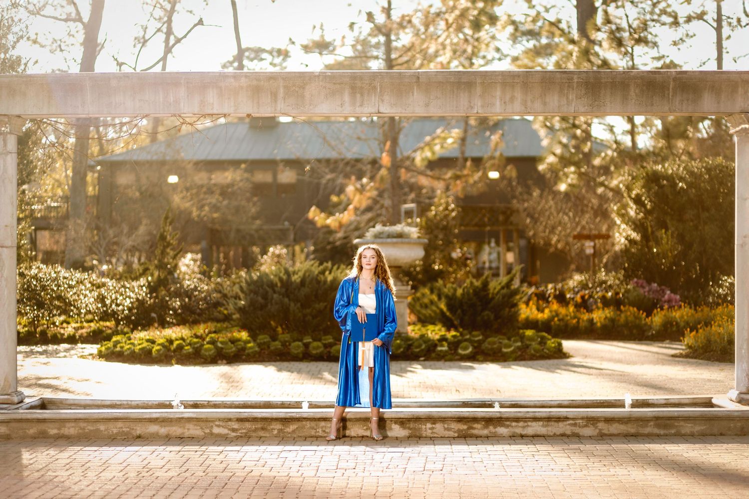 A graduate in a bright blue cap and gown stands in sunlit brick courtyard during golden hour.