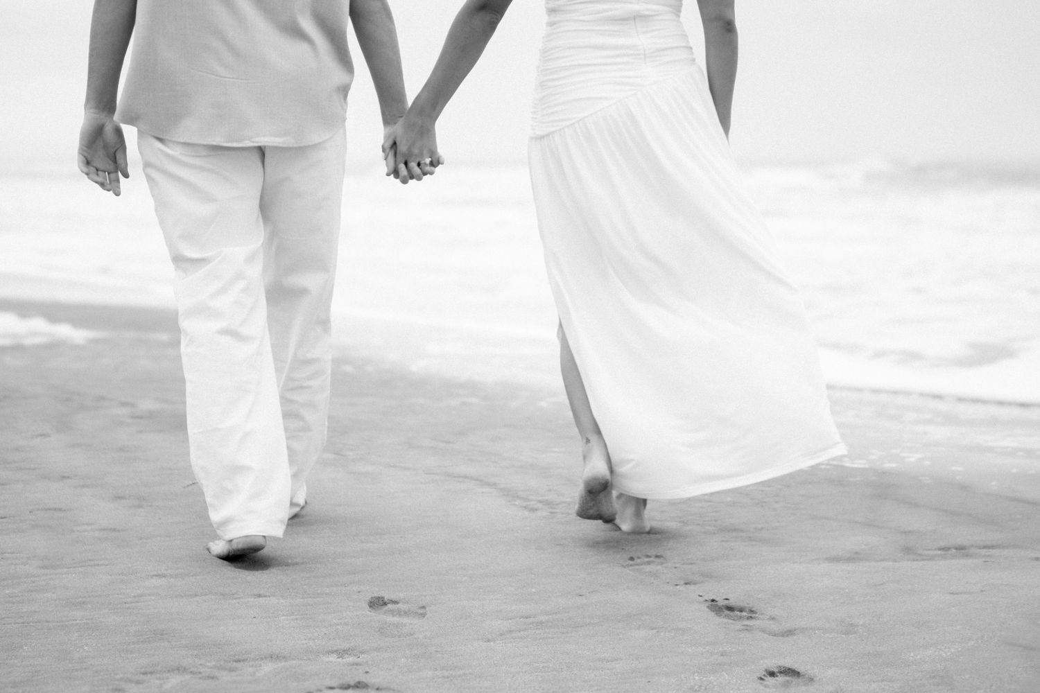 A couple in white attire holds hands while walking along a beach, leaving footprints in the sand.