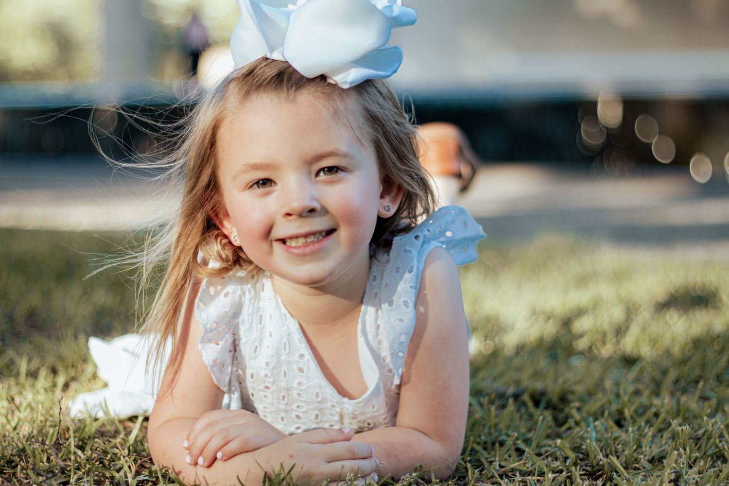 A child in a white lace dress and blue bow lies on grass during golden hour outdoor photo session.