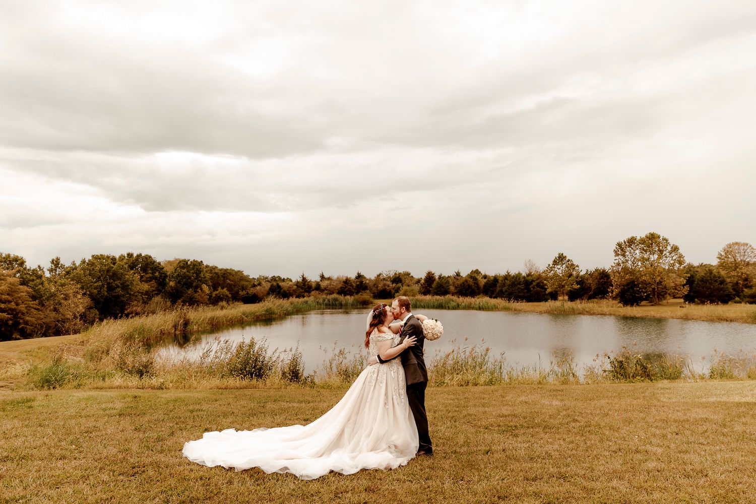 A couple stands on a wooden boardwalk overlooking a serene lake under cloudy skies at a rustic venue.