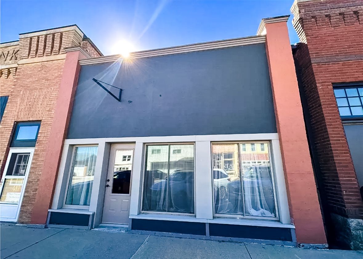 Gray commercial storefront building with large windows and white trim between brick buildings on a sunny day.