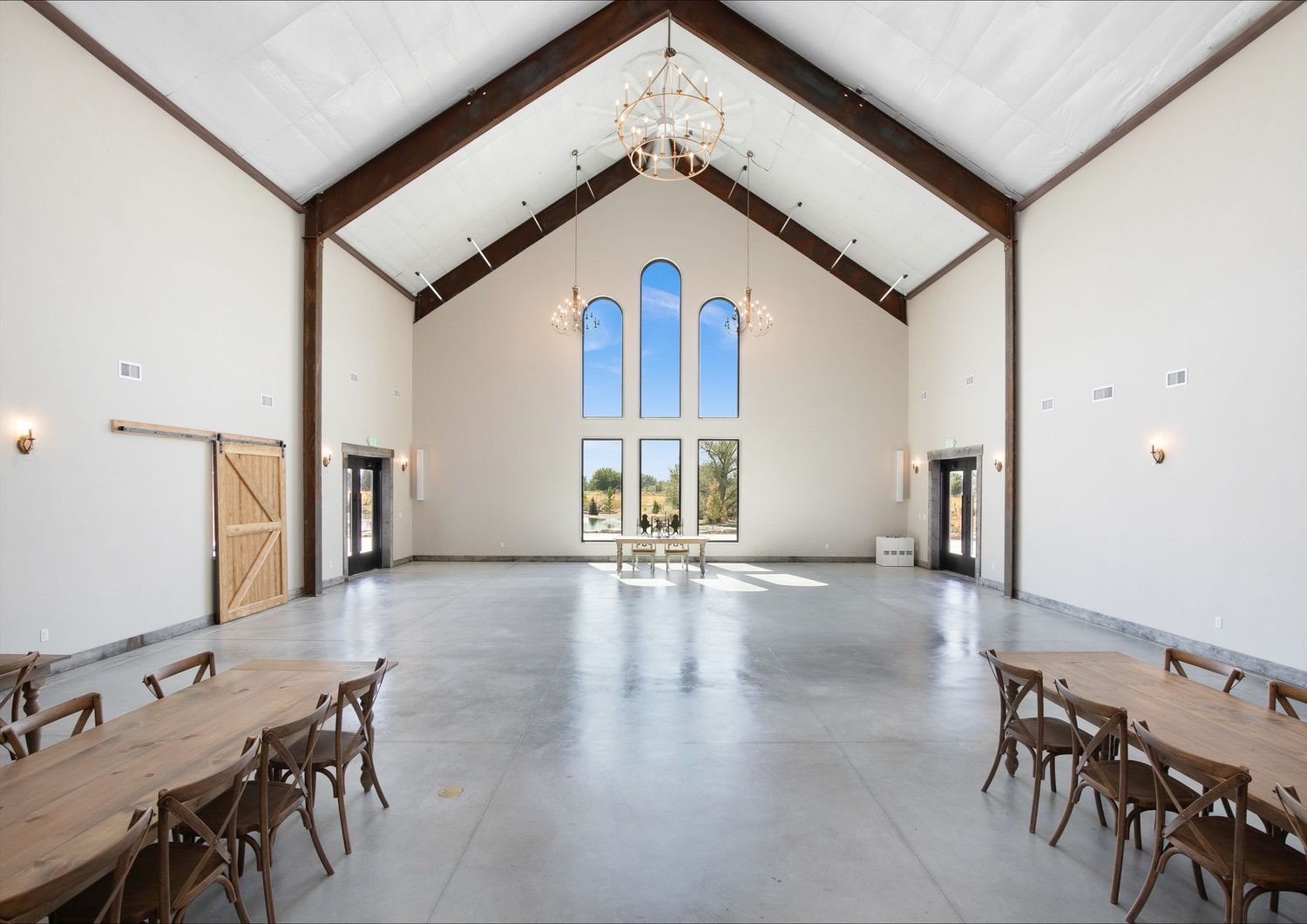 Modern minimalist chapel interior with vaulted ceiling, large arched window, and rustic wooden chairs in rows on polished concrete floor.