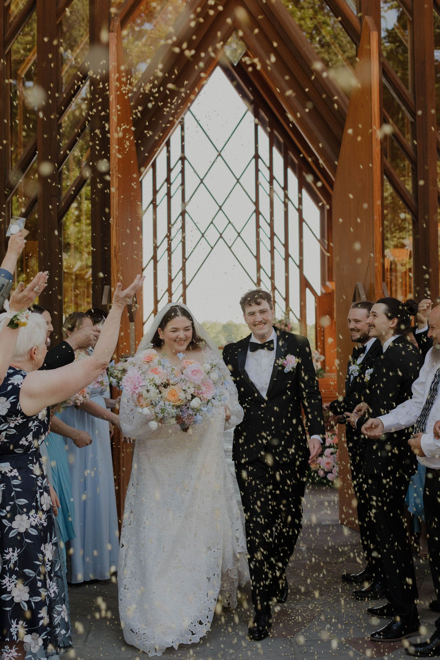 Wedding celebration under a wooden pavilion with guests throwing confetti in joyful celebration.