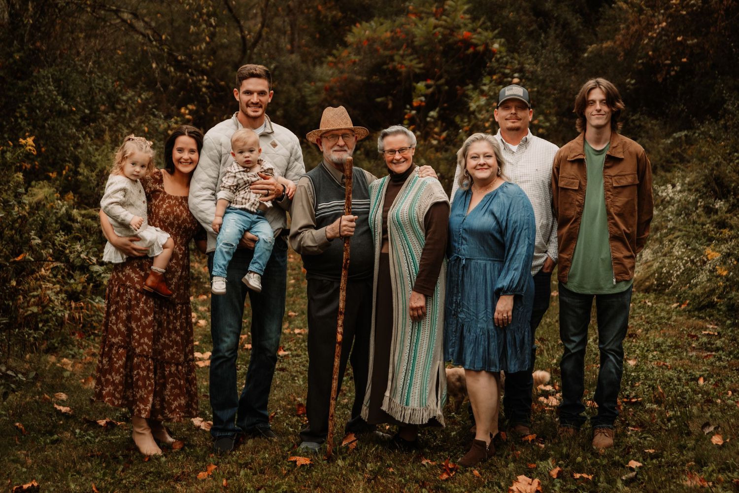 A multi-generational family group standing together outdoors in a wooded setting during autumn.