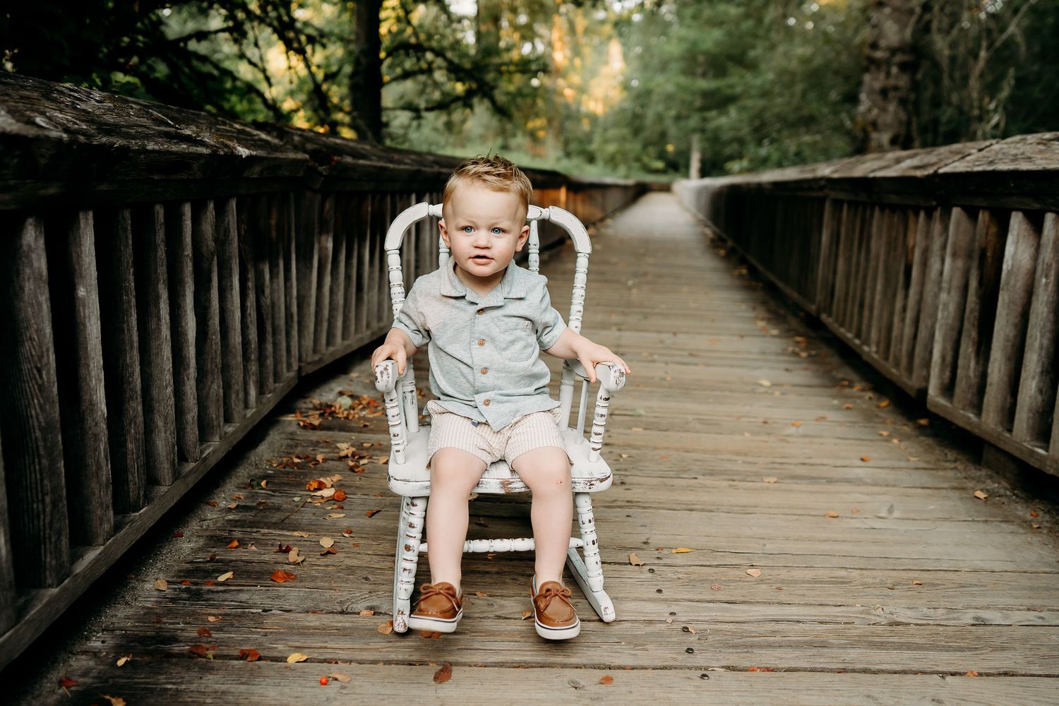 Toddler boy in summer dress clothes sits in a white rocking chair on the boardwalk at Nisqually Wildlife Refuge for summer birthday photos.