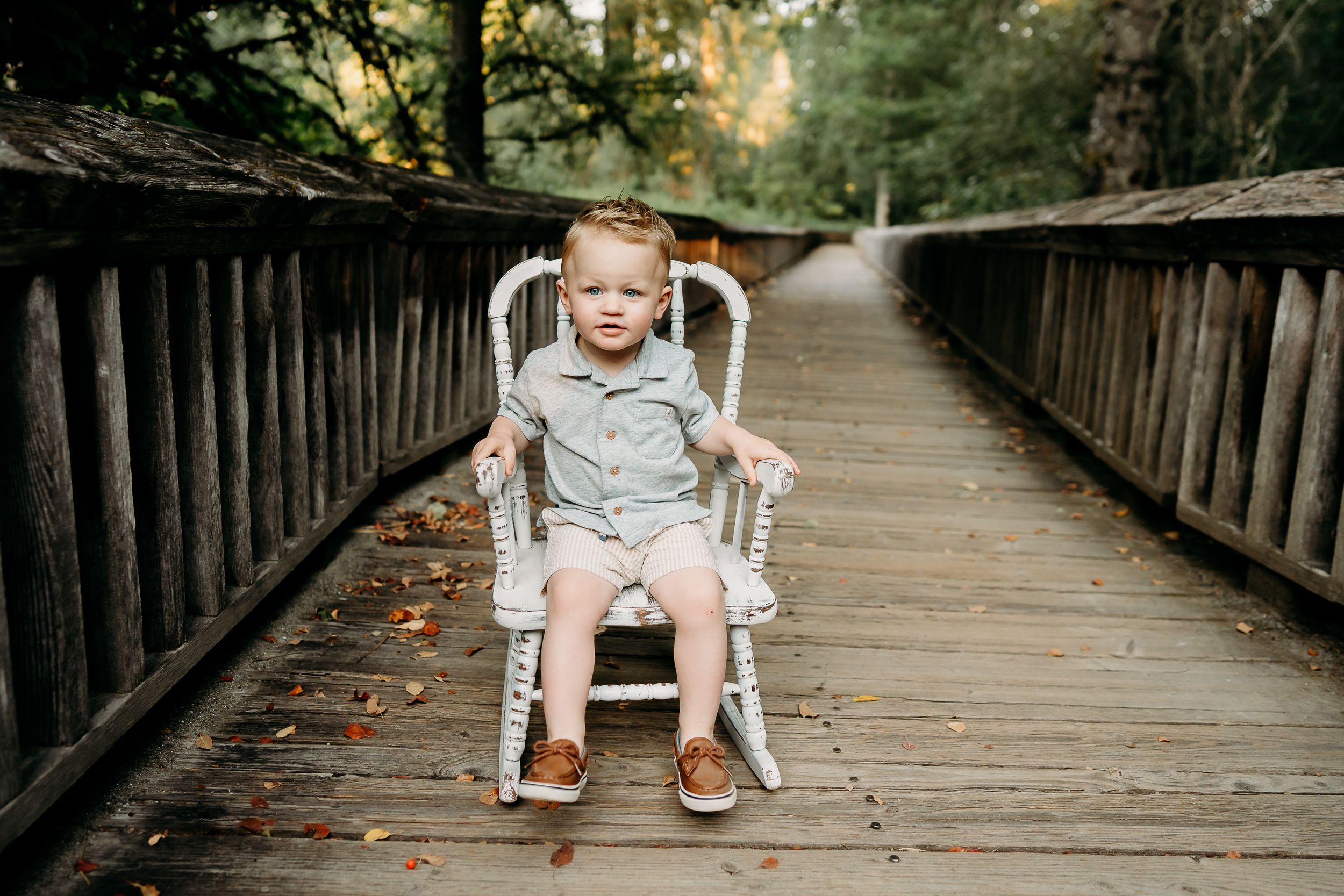 Toddler boy in summer dress clothes sits in a white rocking chair on the boardwalk at Nisqually Wildlife Refuge for summer birthday photos.