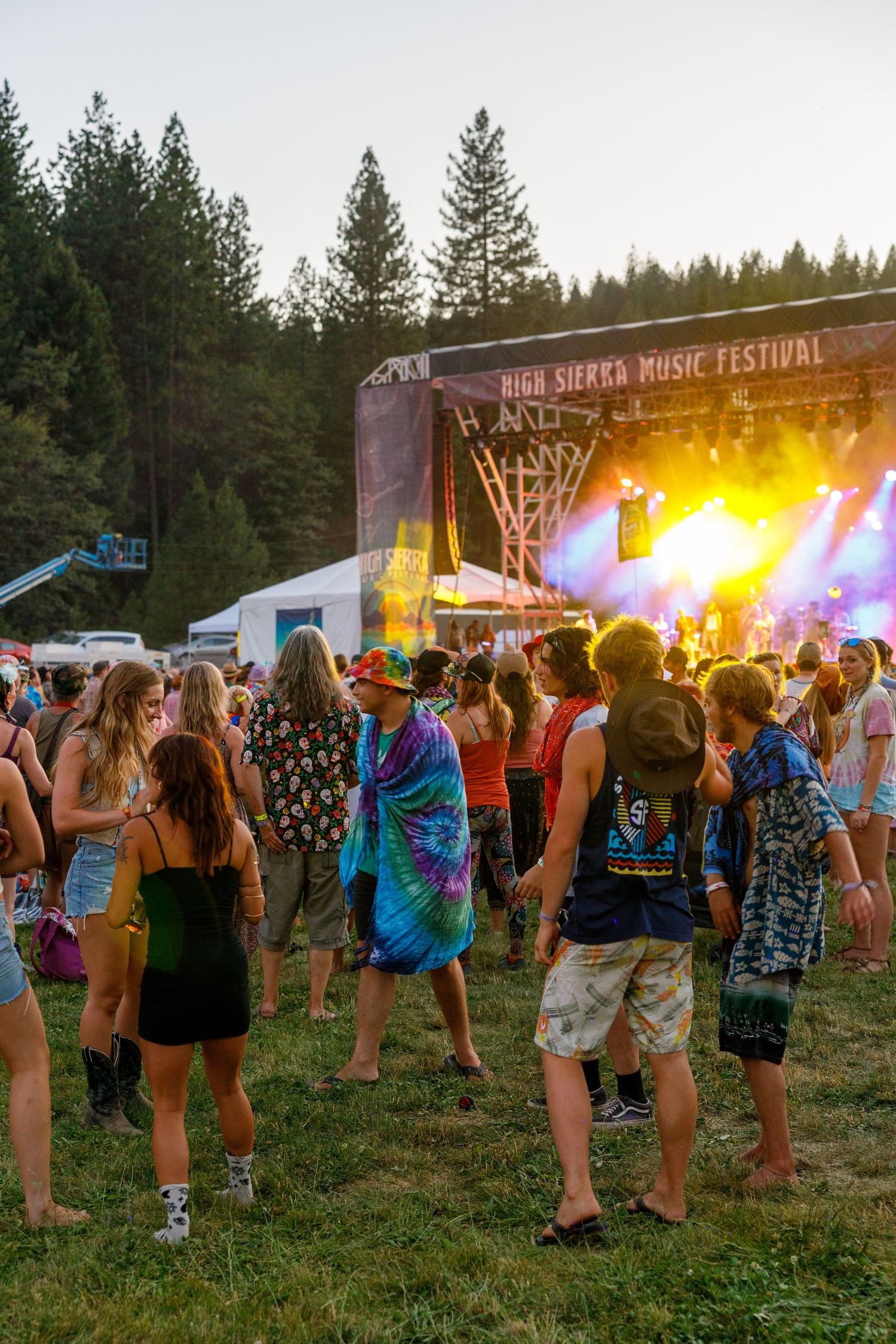 Crowd gathers at outdoor music festival stage with sunset lighting and pine trees in background.
