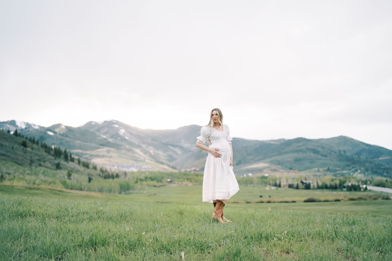 A person in a white dress stands barefoot in a green meadow with rolling mountains in the background.