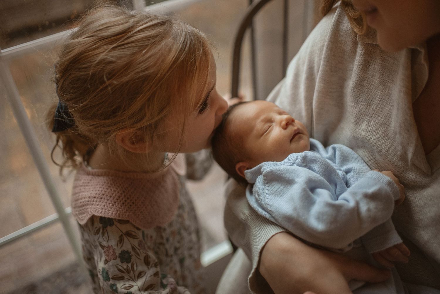A tender moment between siblings as little girl gives a gentle kiss to baby in light blue sleeper.