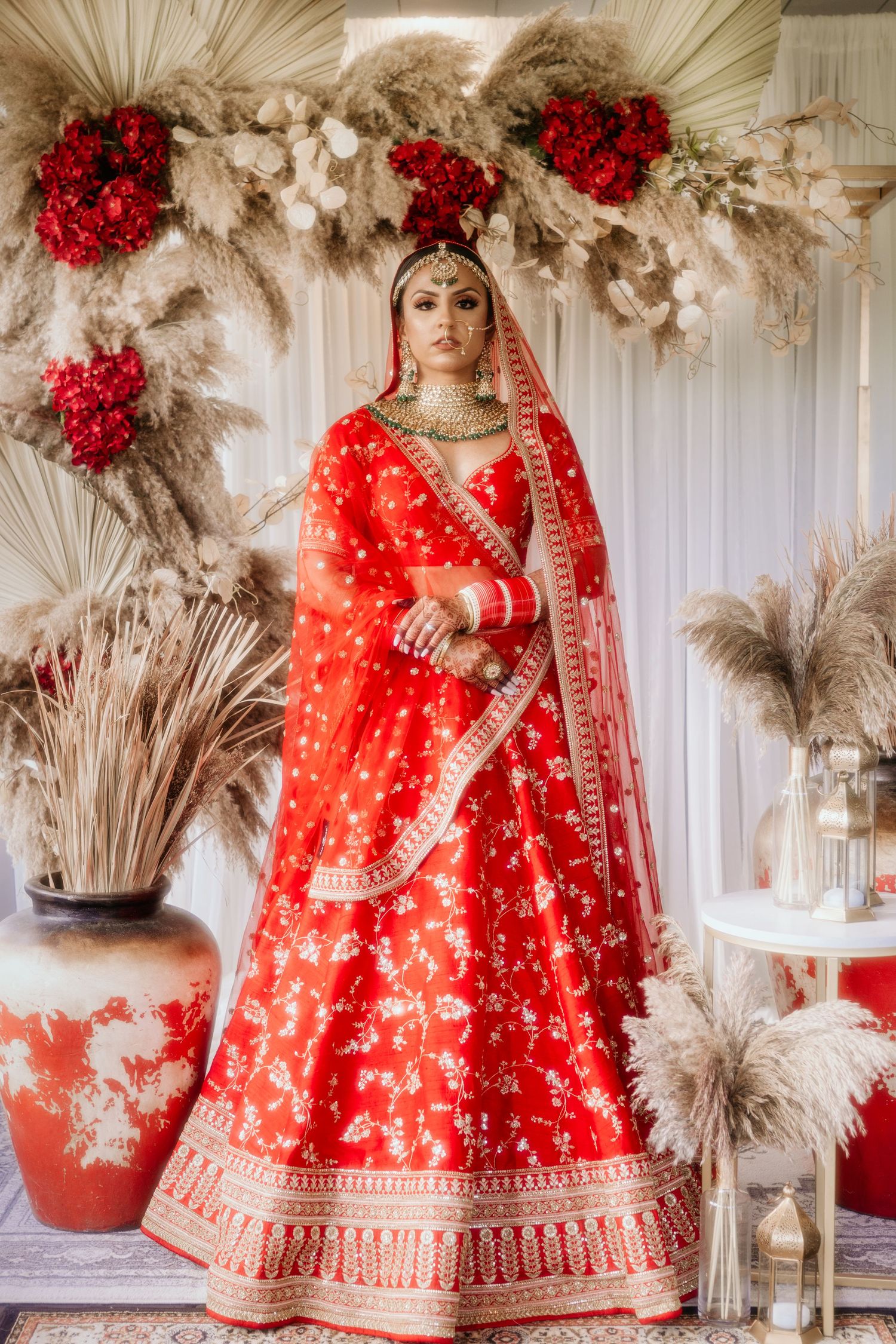Traditional bride wearing an ornate red bridal lehenga poses against a decorative backdrop with red flowers and pampas grass.
