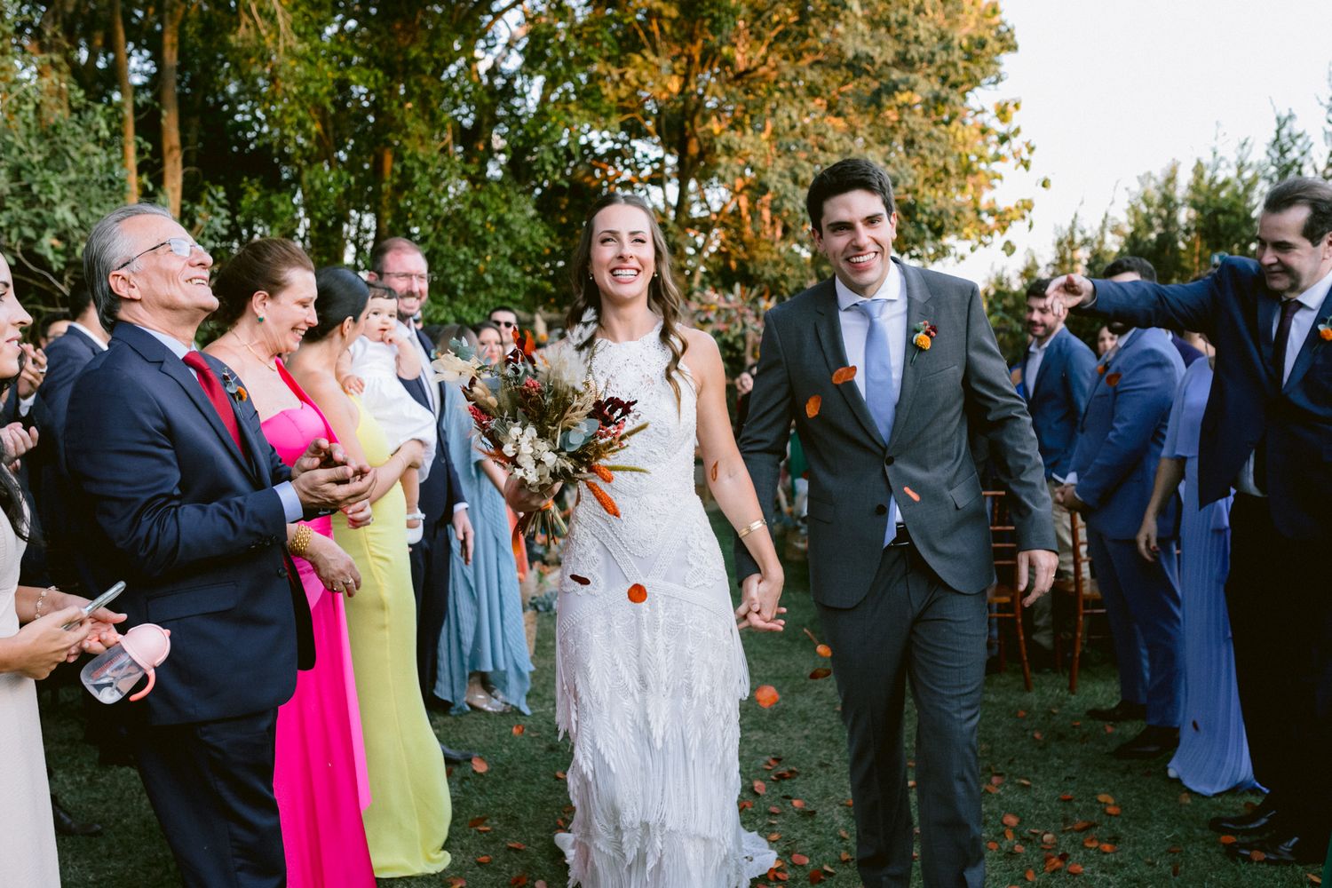 Wedding guests celebrate by tossing autumn leaves into the air while standing on grass during an outdoor ceremony.