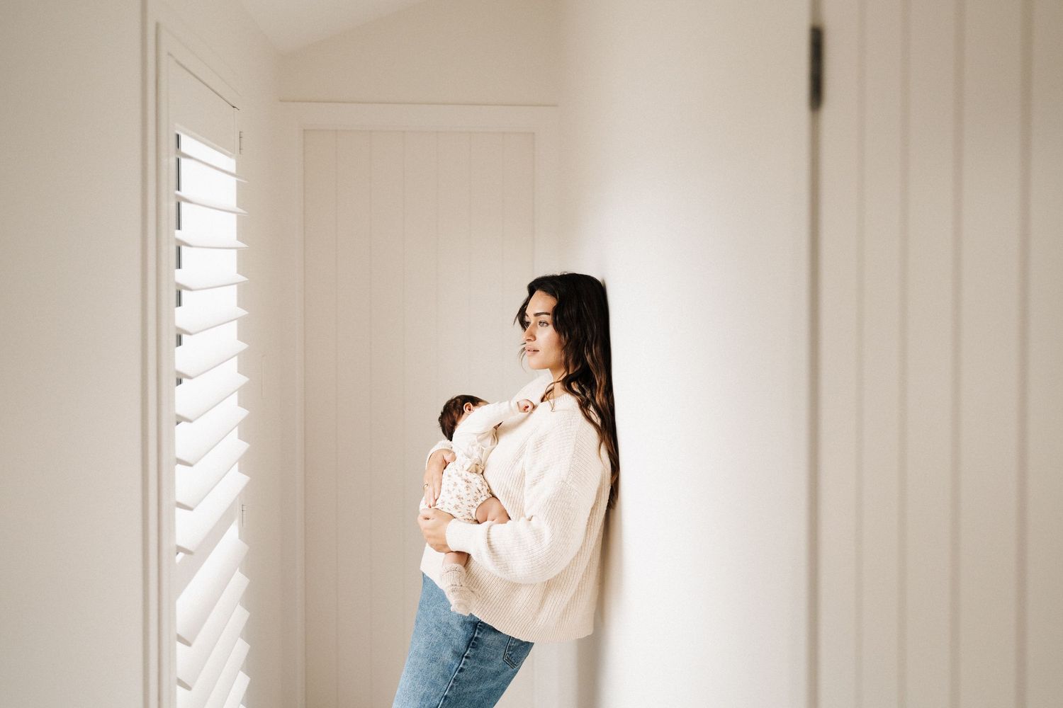 Side profile of parent in pale sweater holding infant near bright window with plantation shutters.