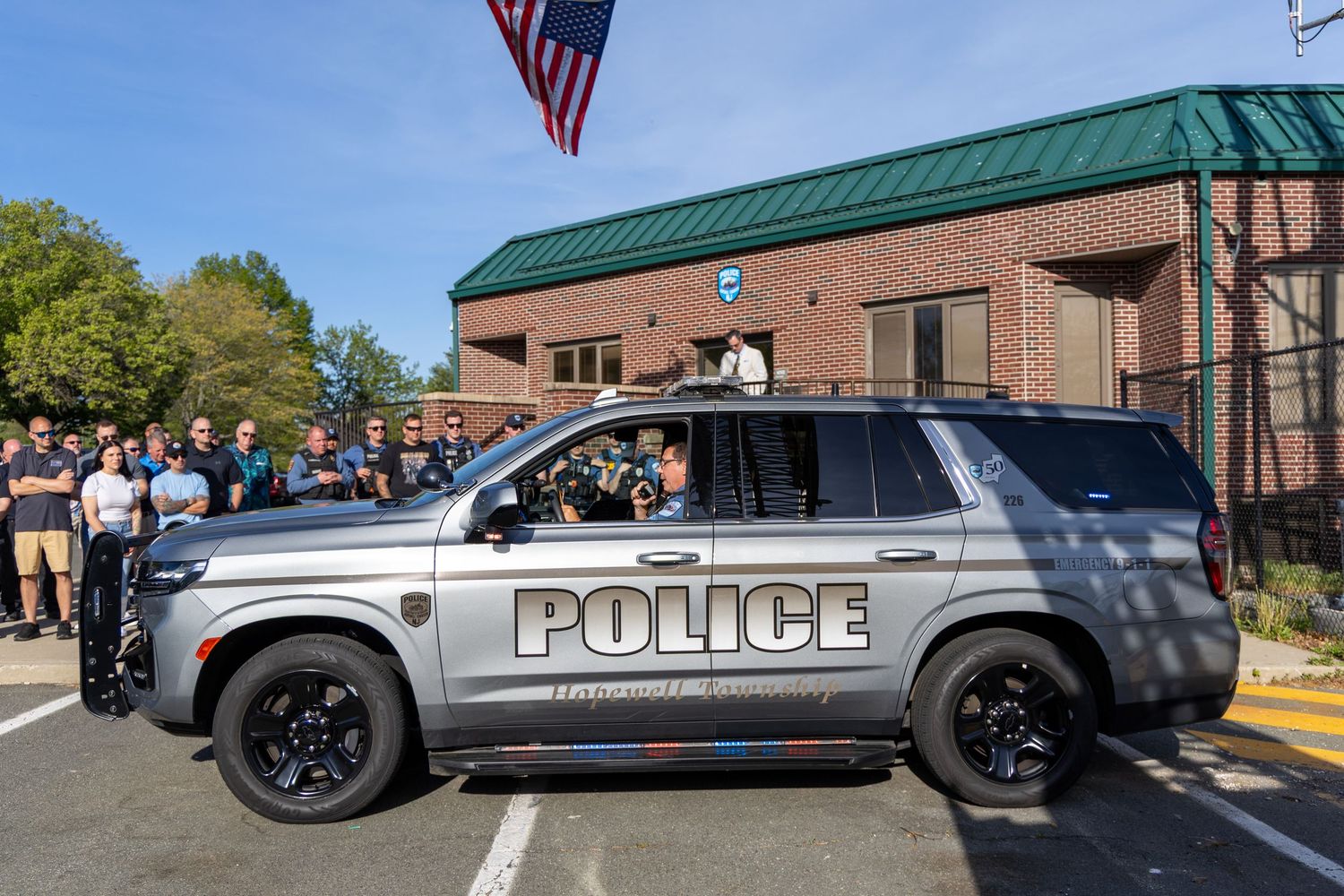 A silver police SUV vehicle is parked outside a building with a green roof and American flag flying above.