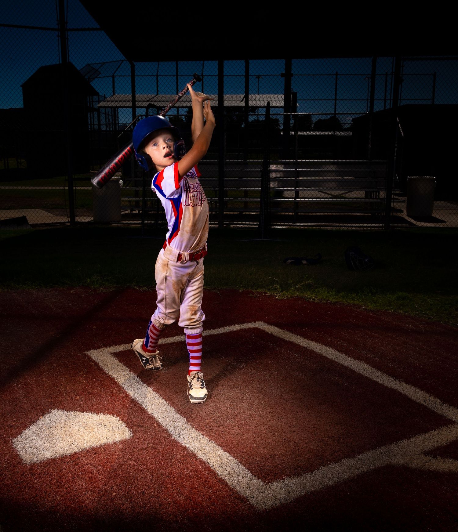 Young baseball player practices batting stance in dramatic lighting on home plate.