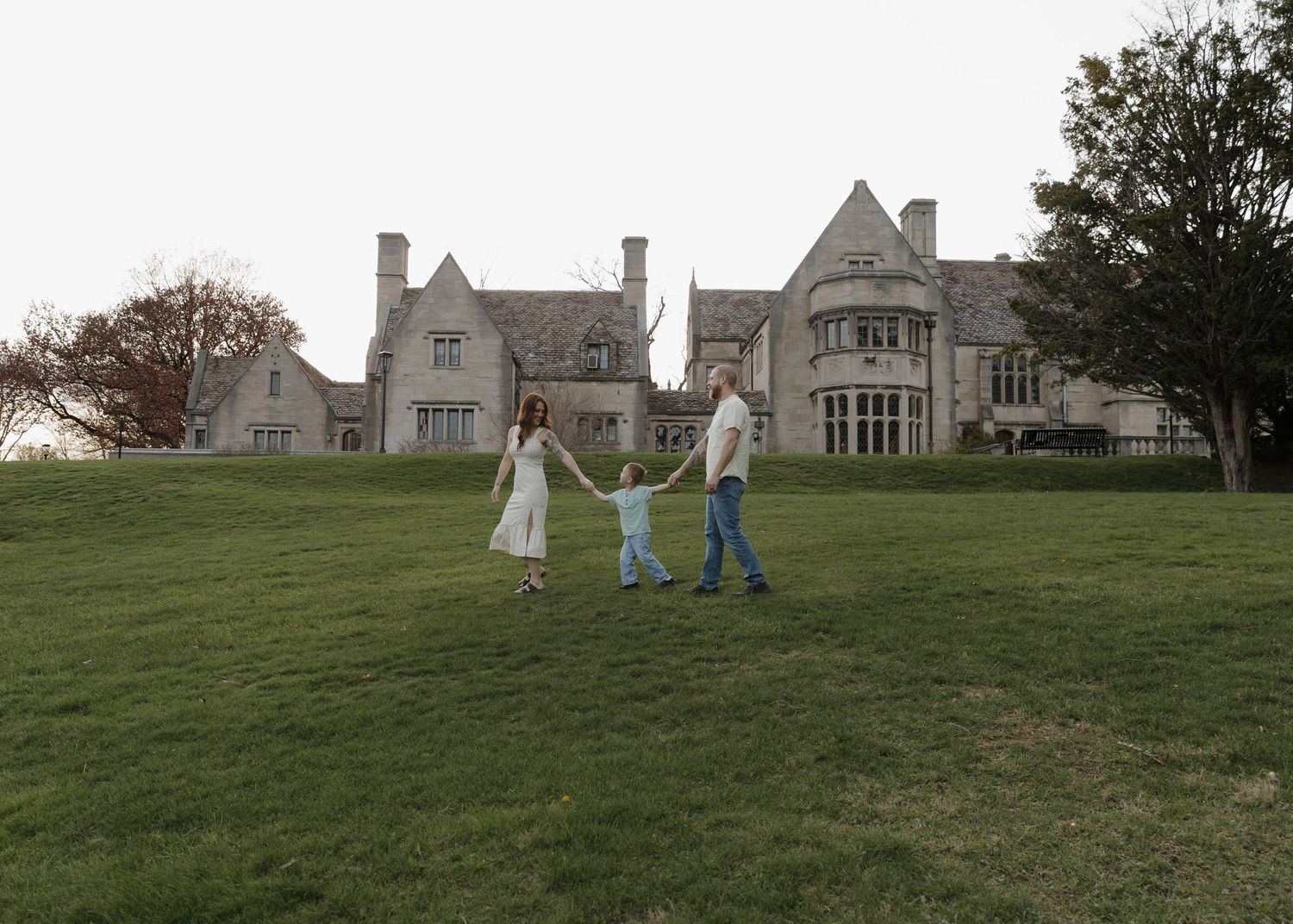 Three people hold hands while walking on a green lawn in front of a grand stone mansion on an overcast day.