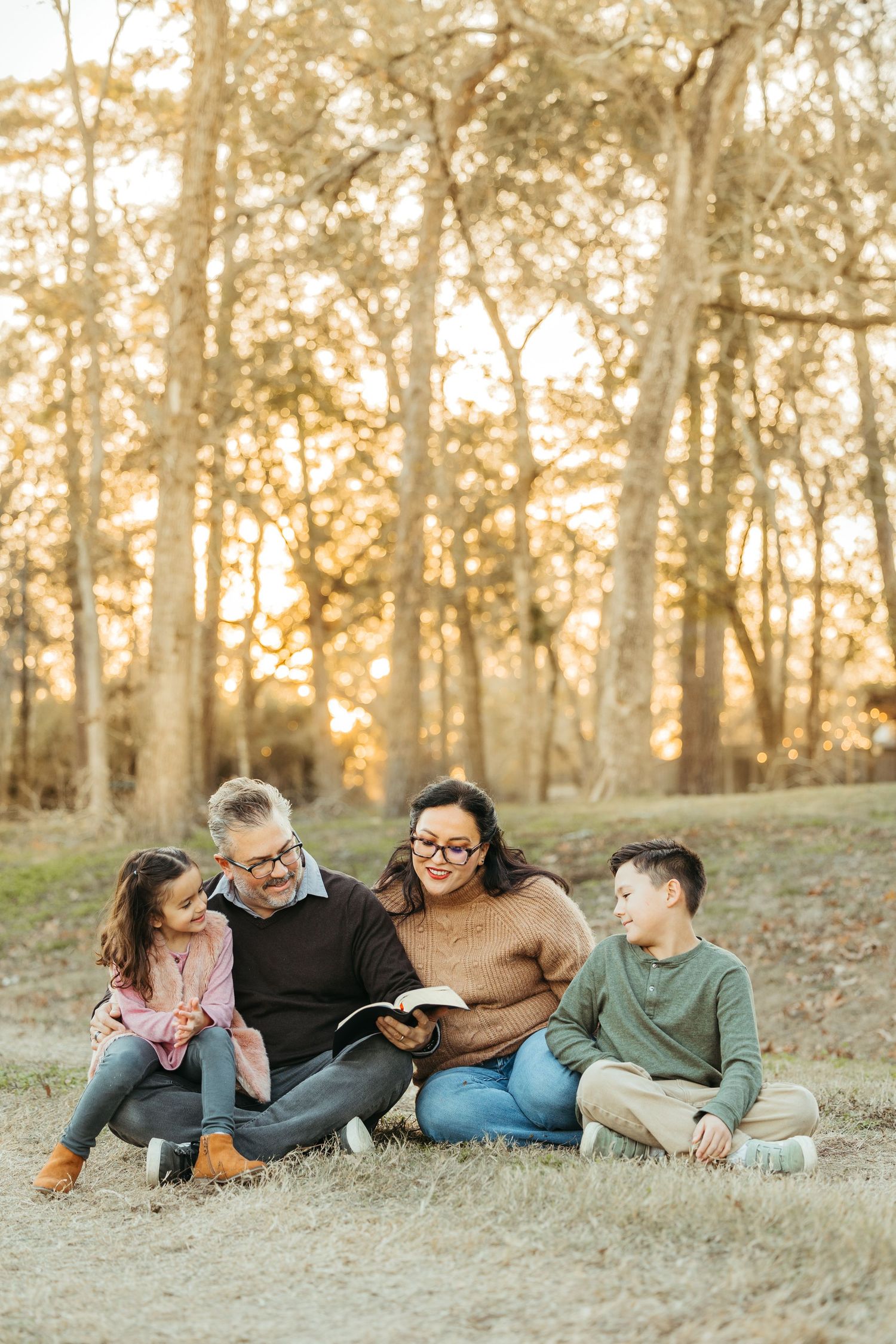 A family of four sits together on grass in a golden forest, reading a book and smiling.