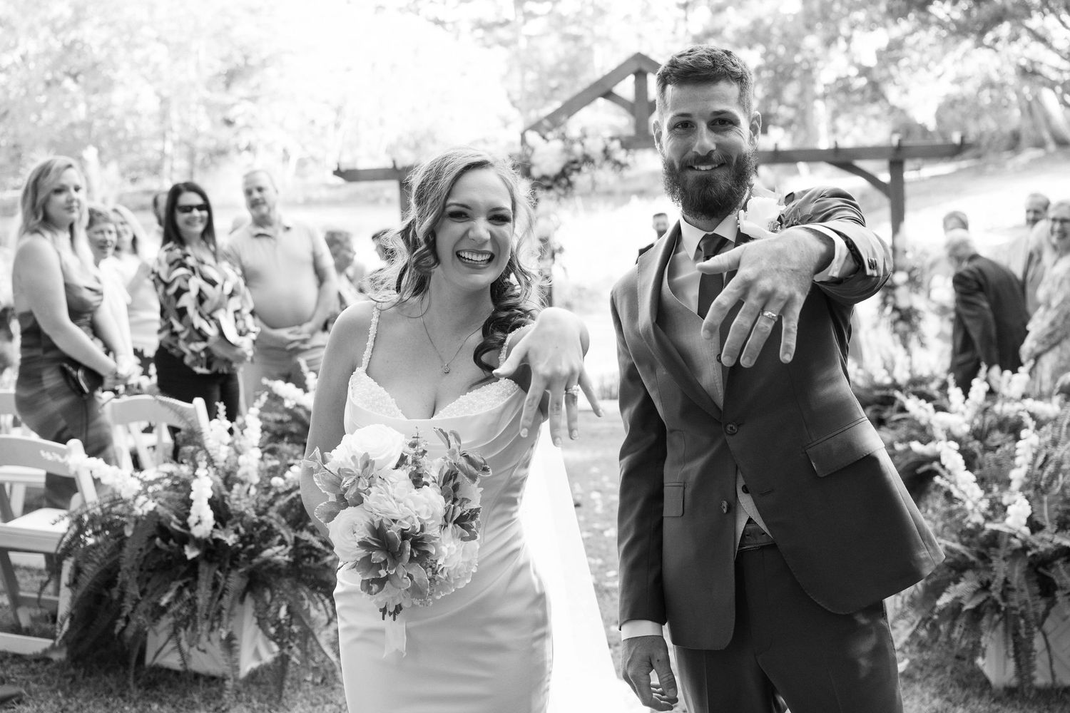 Smiling bride in a white dress and groom in a gray suit laugh while leaning against a wooden post at an outdoor forest wedding.