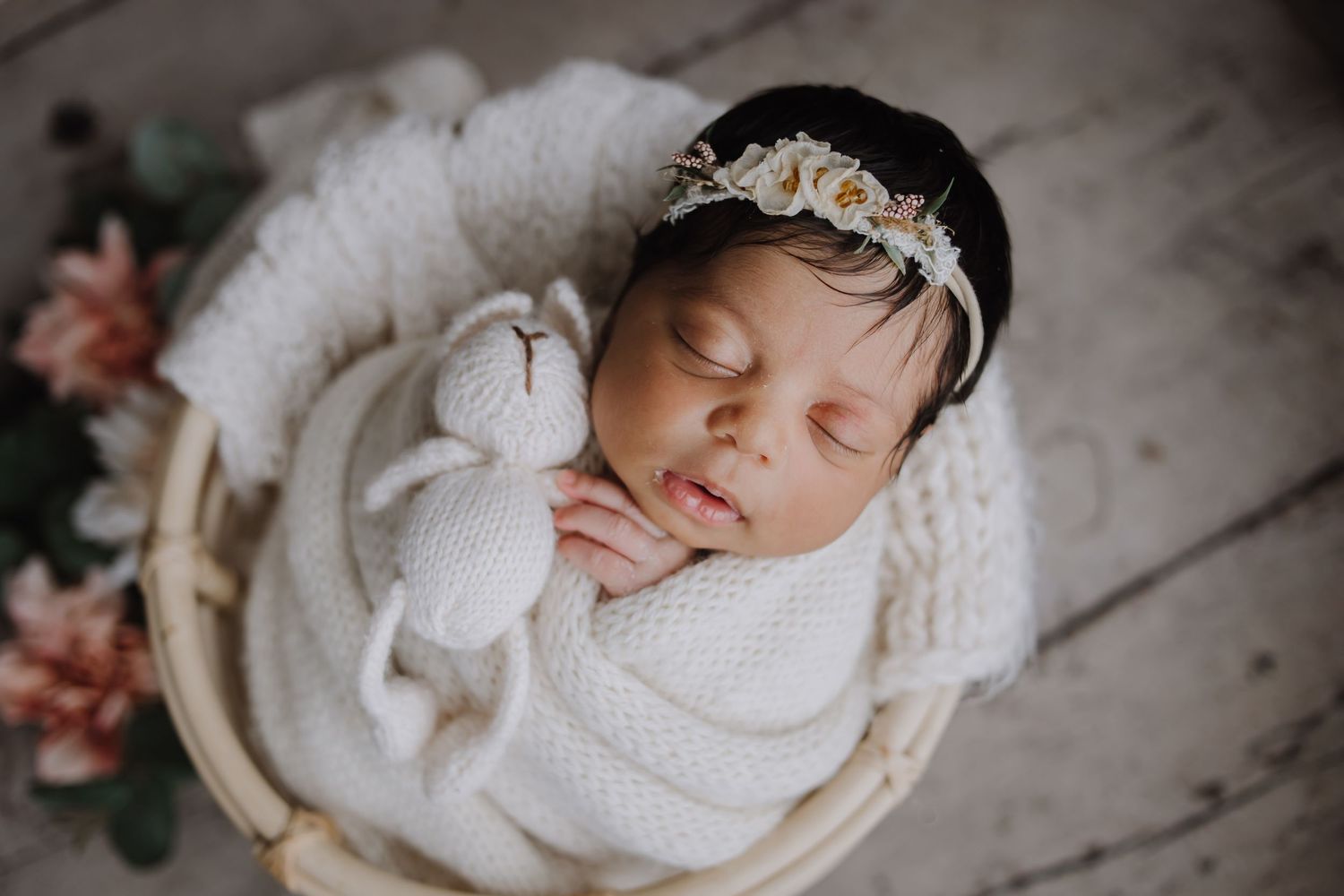 Sleeping newborn baby wrapped in white knit blanket holding stuffed bunny in wicker basket with floral headband.
