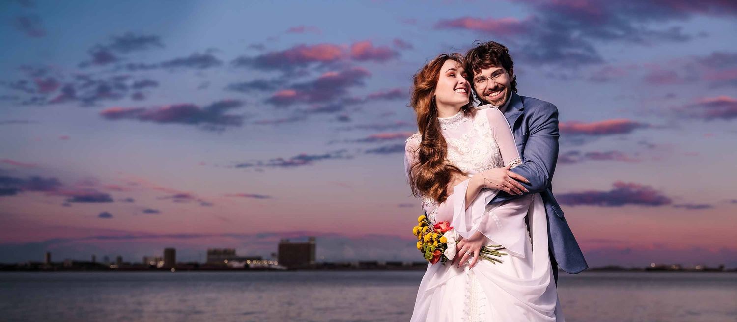 A couple in wedding attire embraces at sunset Fan Pier Park in Boston, the bride holding yellow flowers under a colorful sky.