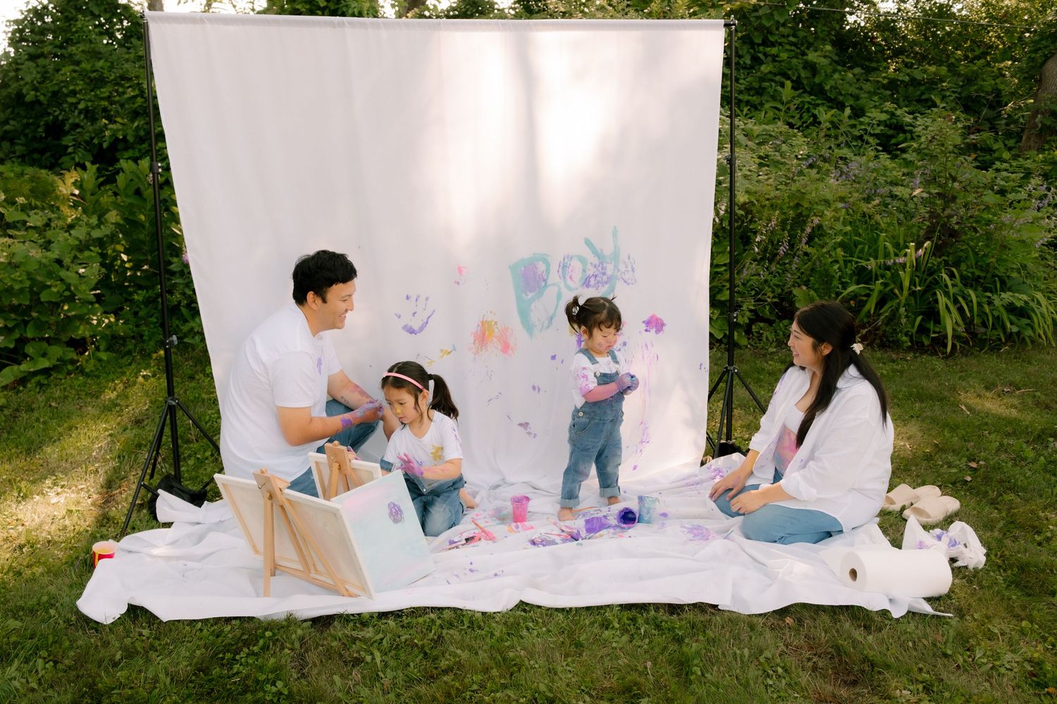 Family of four enjoying an outdoor painting activity on a white blanket with a backdrop in a lush green garden.