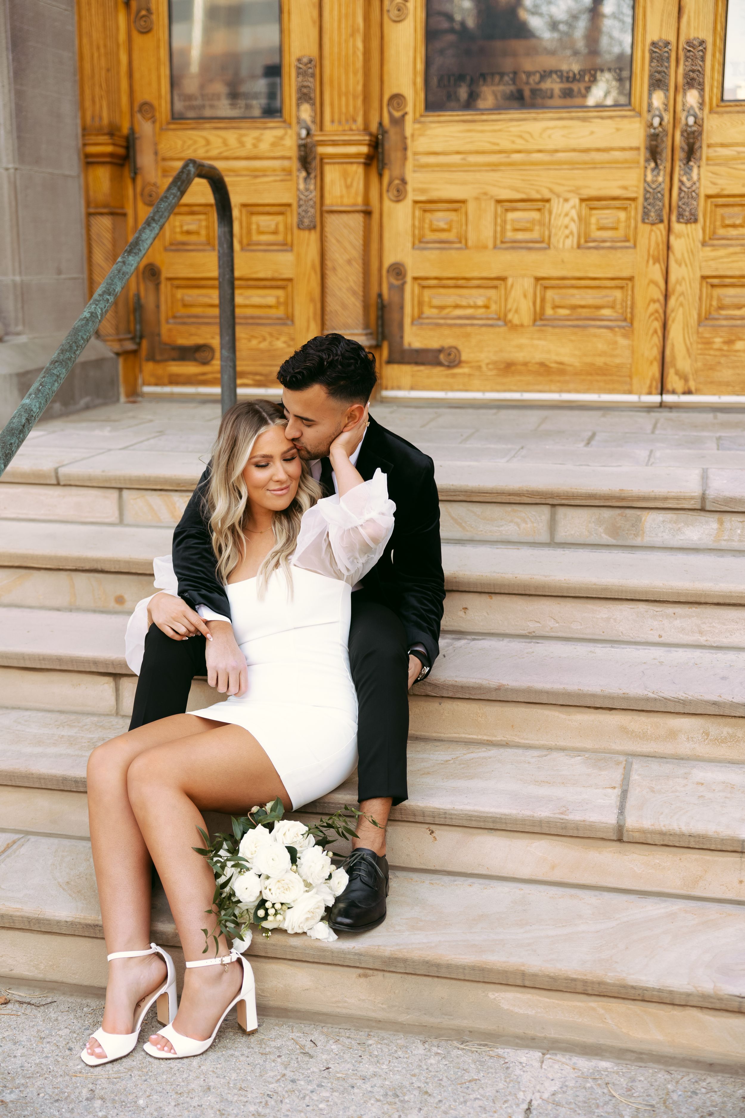 Couple in chic engagement picture outfits sitting on city hall steps. Man in formal suit. Woman in white mini dress.