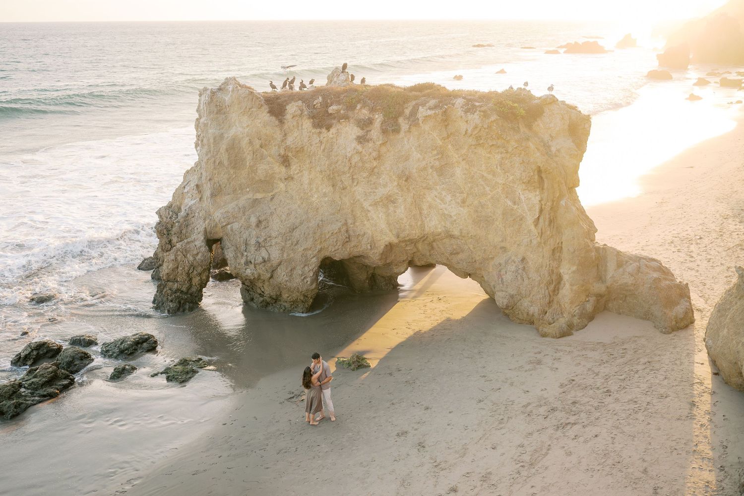 Couple embraces under a natural rock arch on a sandy beach at golden sunset in Malibu, California.