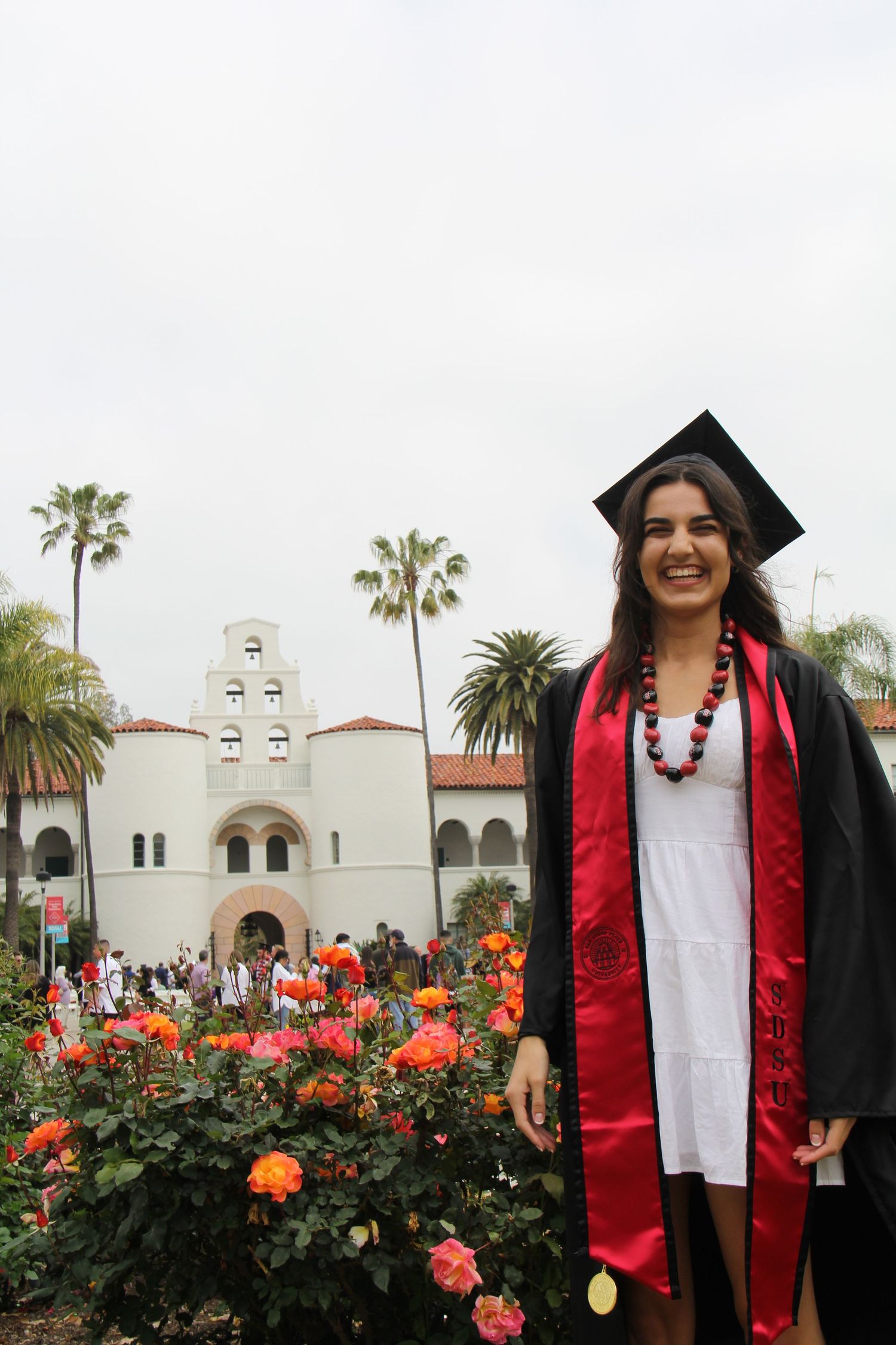 Graduate in red and black commencement regalia poses in front of Spanish-style building with rose garden.