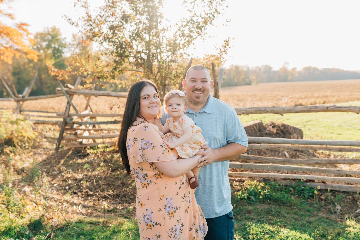 A mother holds her 2 year old daughter while her husband snugs close while standing backlit on a open field.