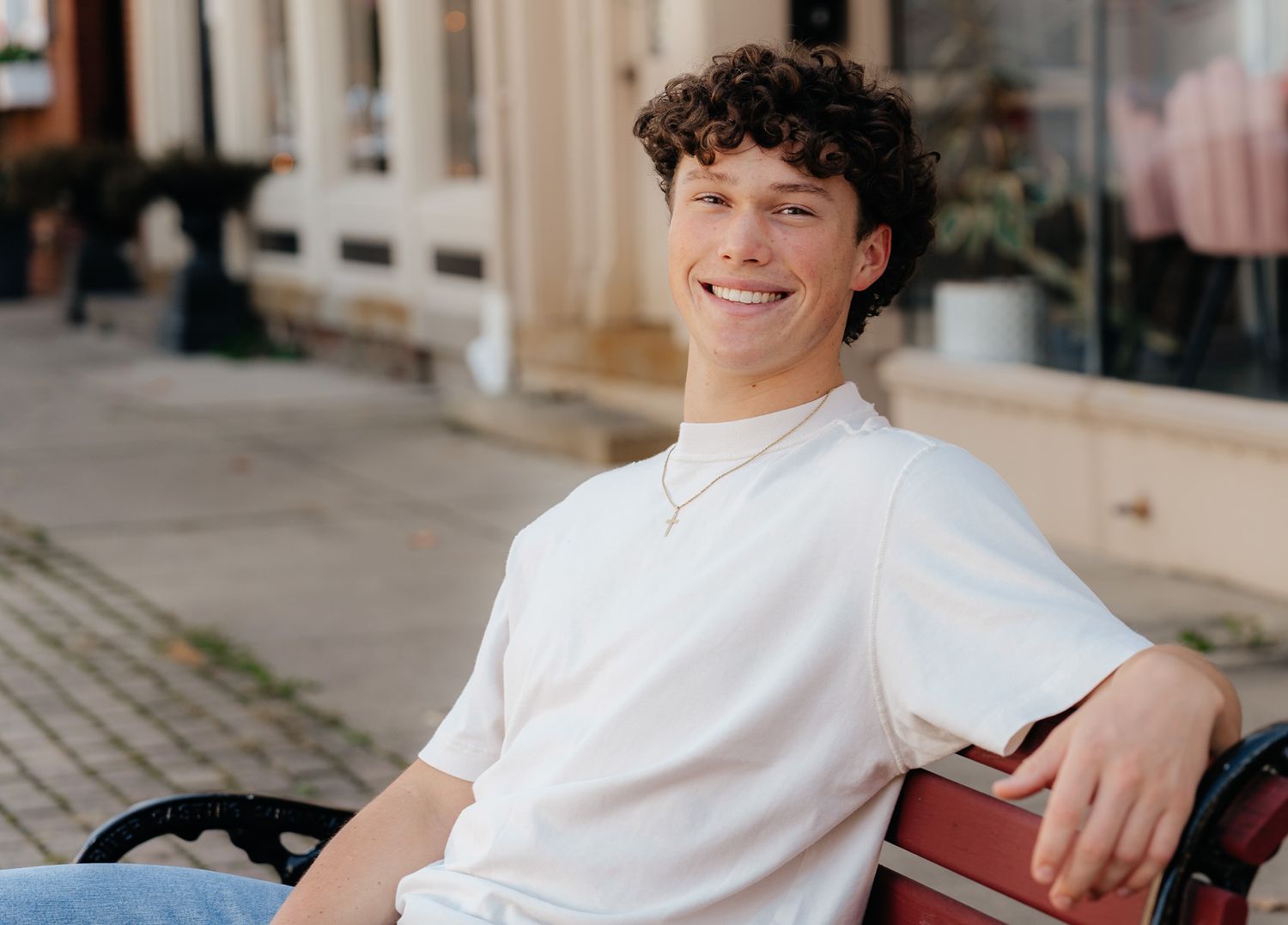 A person in a white t-shirt sits casually on a bench outside storefronts on a cobblestone street.